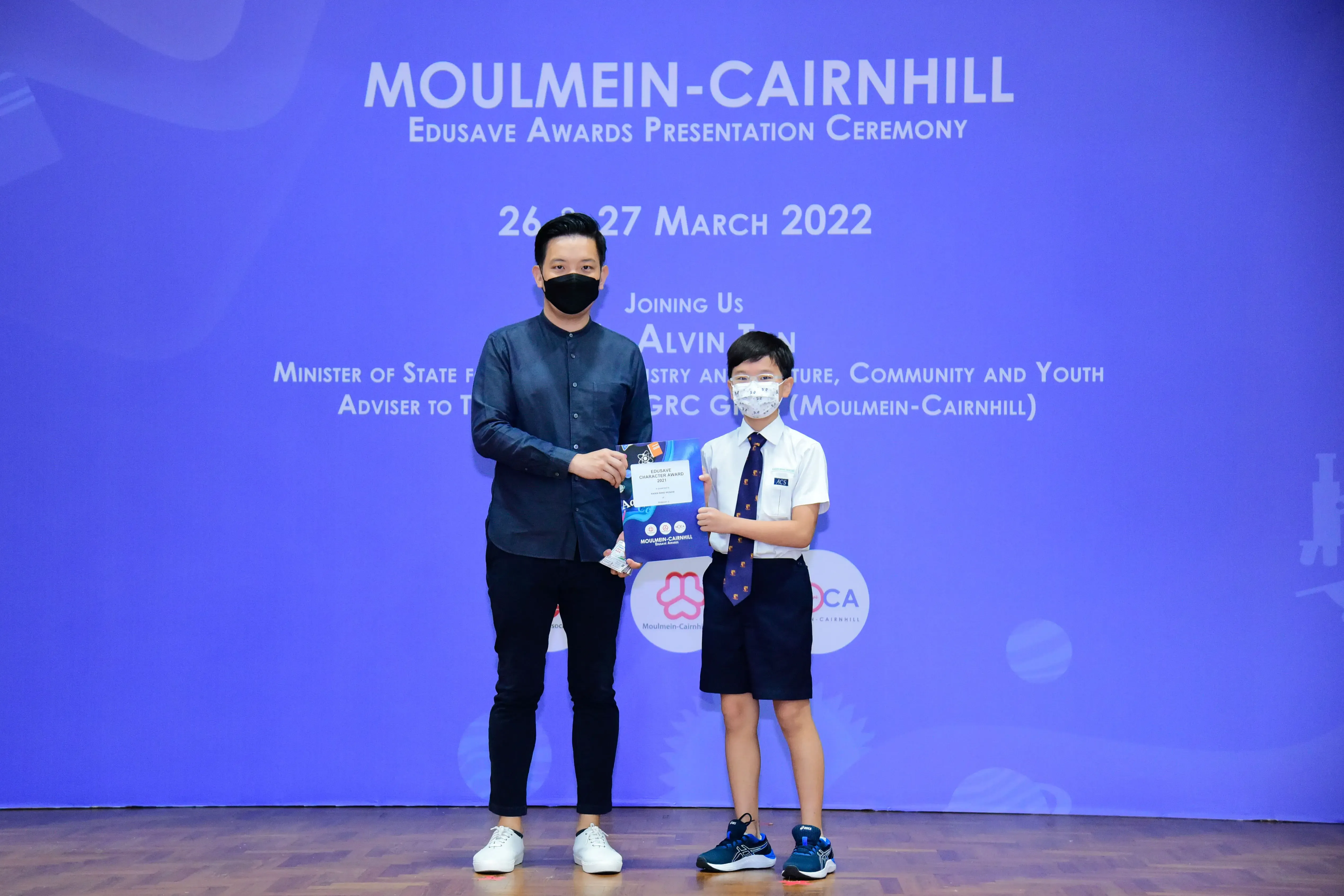 An adult and a young boy in school uniform hold a certificate together at the Moulmein-Cairnhill Edusave Awards Ceremony 2022.