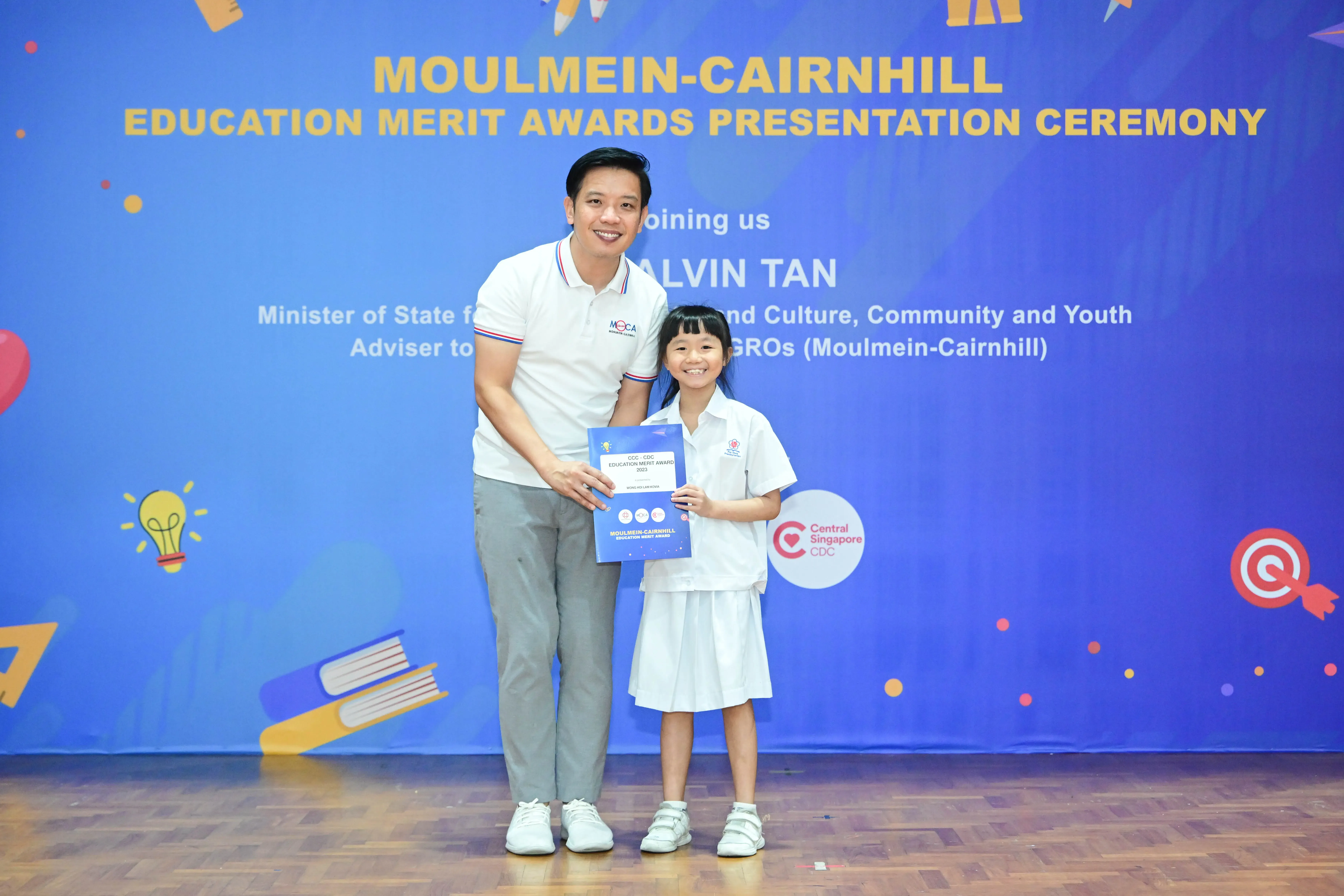 A man and a young girl in school uniform hold an award certificate at the Moulmein-Cairnhill Education Merit Awards ceremony.