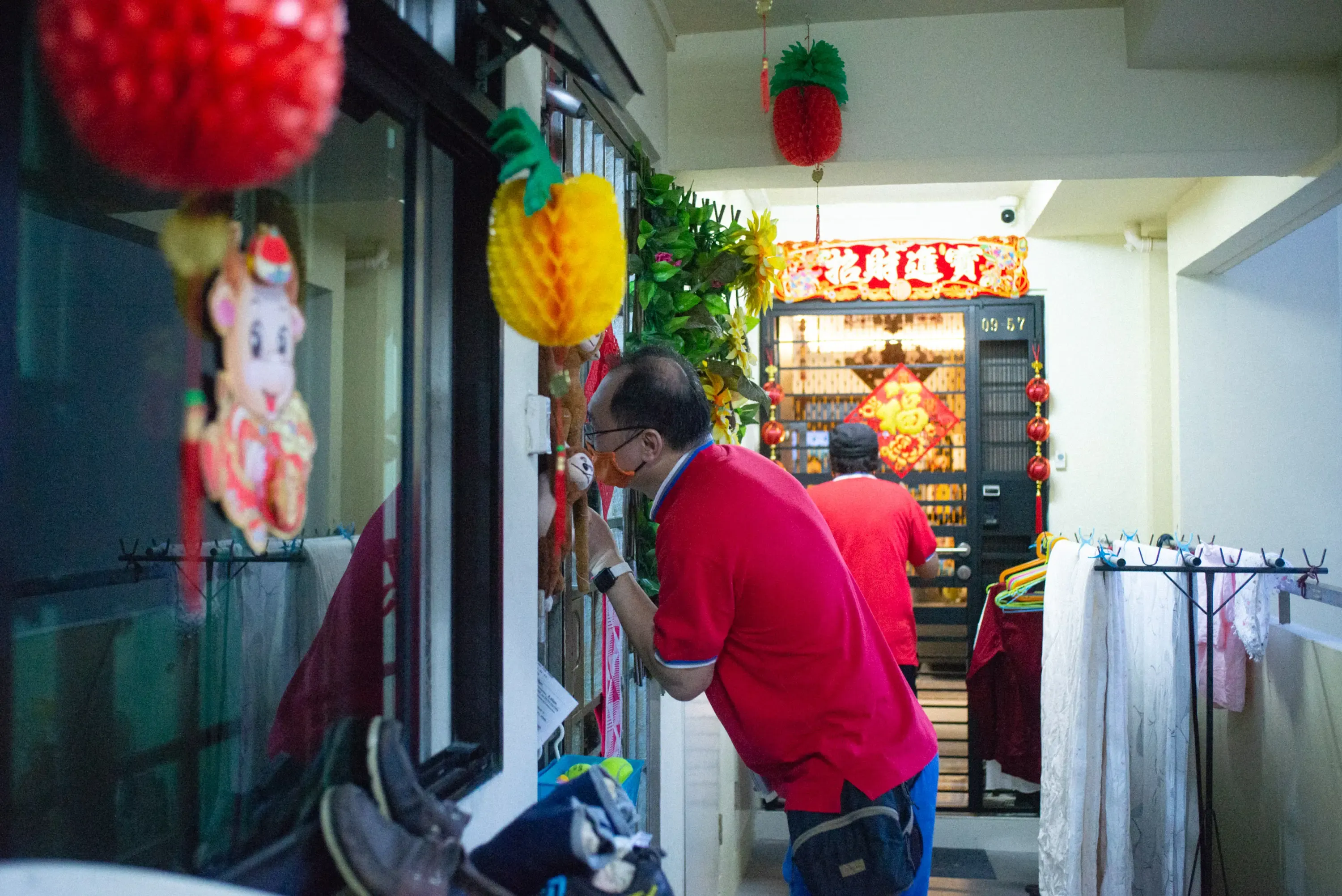 Man in red shirt bending near a gate decorated with Chinese New Year ornaments including pineapple and red decorations.