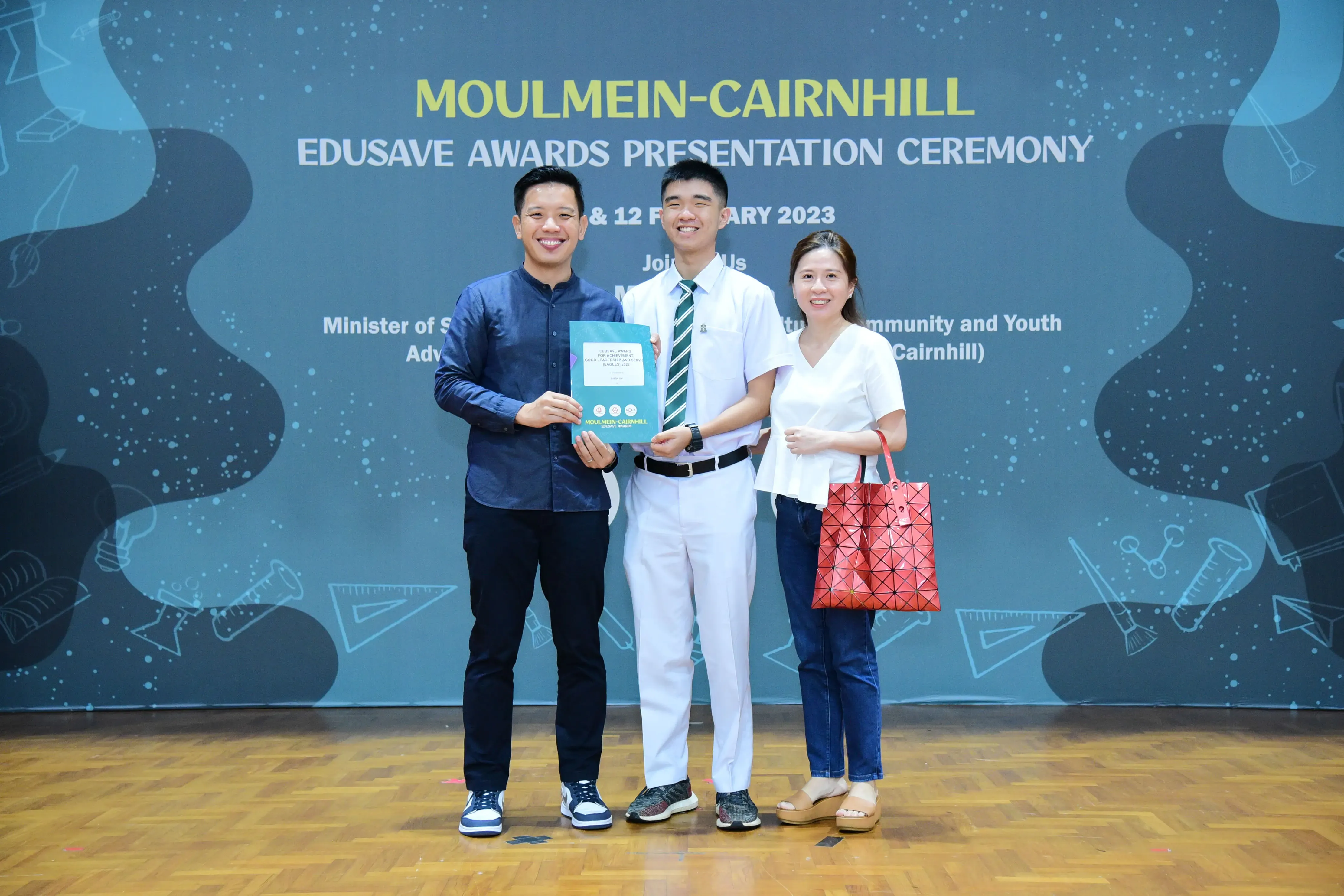 Three people smile holding an award booklet at the Moulmein-Cairnhill Edusave Awards Presentation Ceremony 2023.