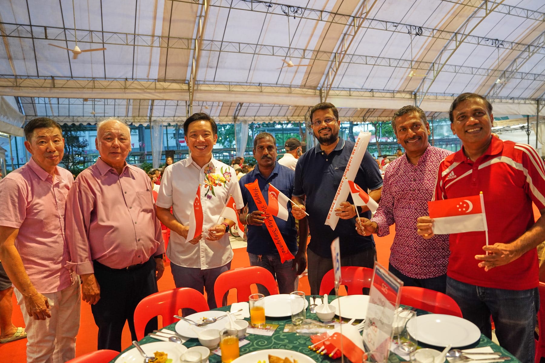 Six men smile and hold Singapore flags at a festive outdoor dinner event under a large tent.