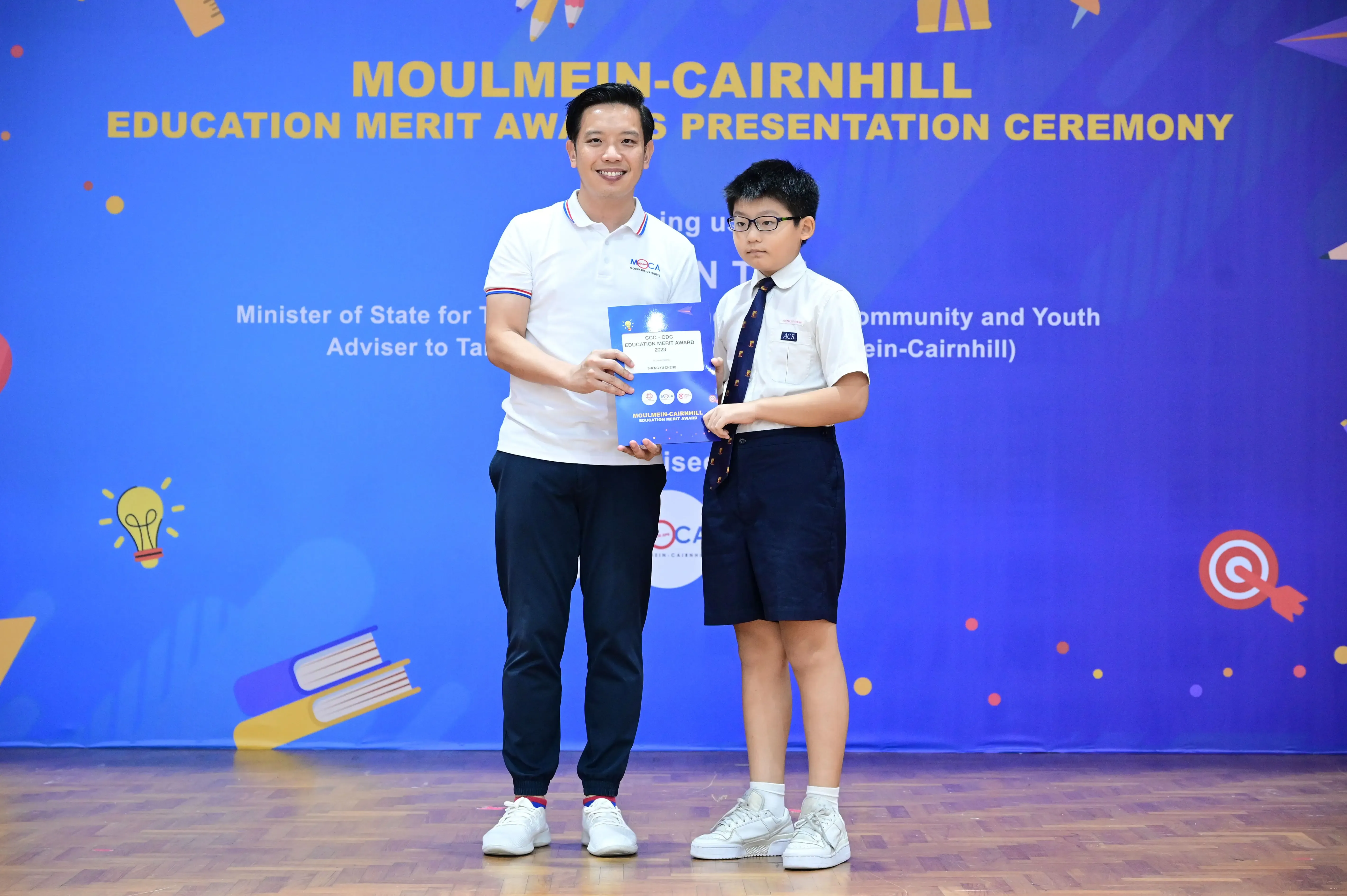 An adult and a young boy in school uniform hold an award certificate at the Moulmein-Cairnhill Education Merit Awards ceremony.