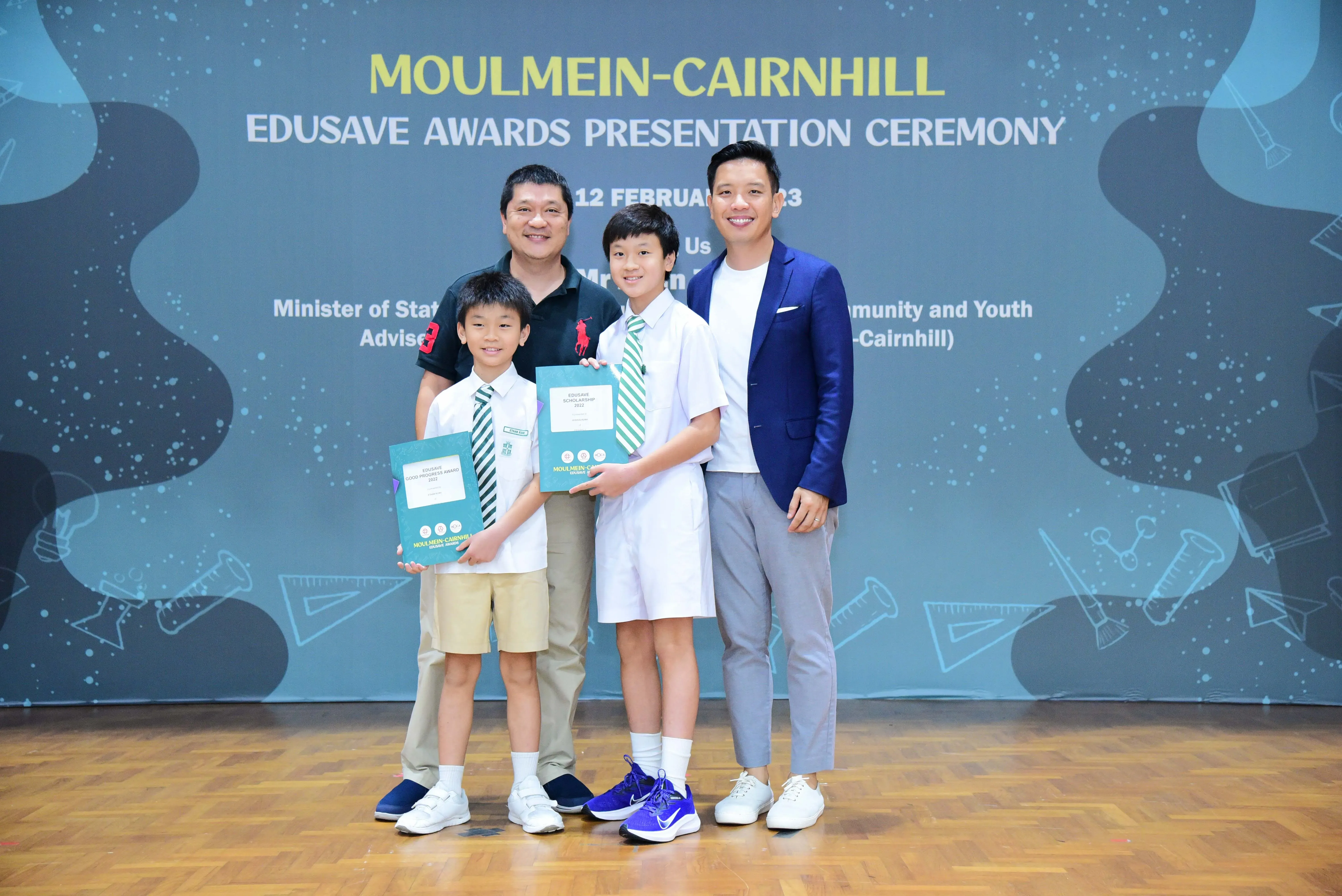 Two boys holding award certificates pose with two men at the Moulmein-Cairnhill Edusave Awards Presentation Ceremony.
