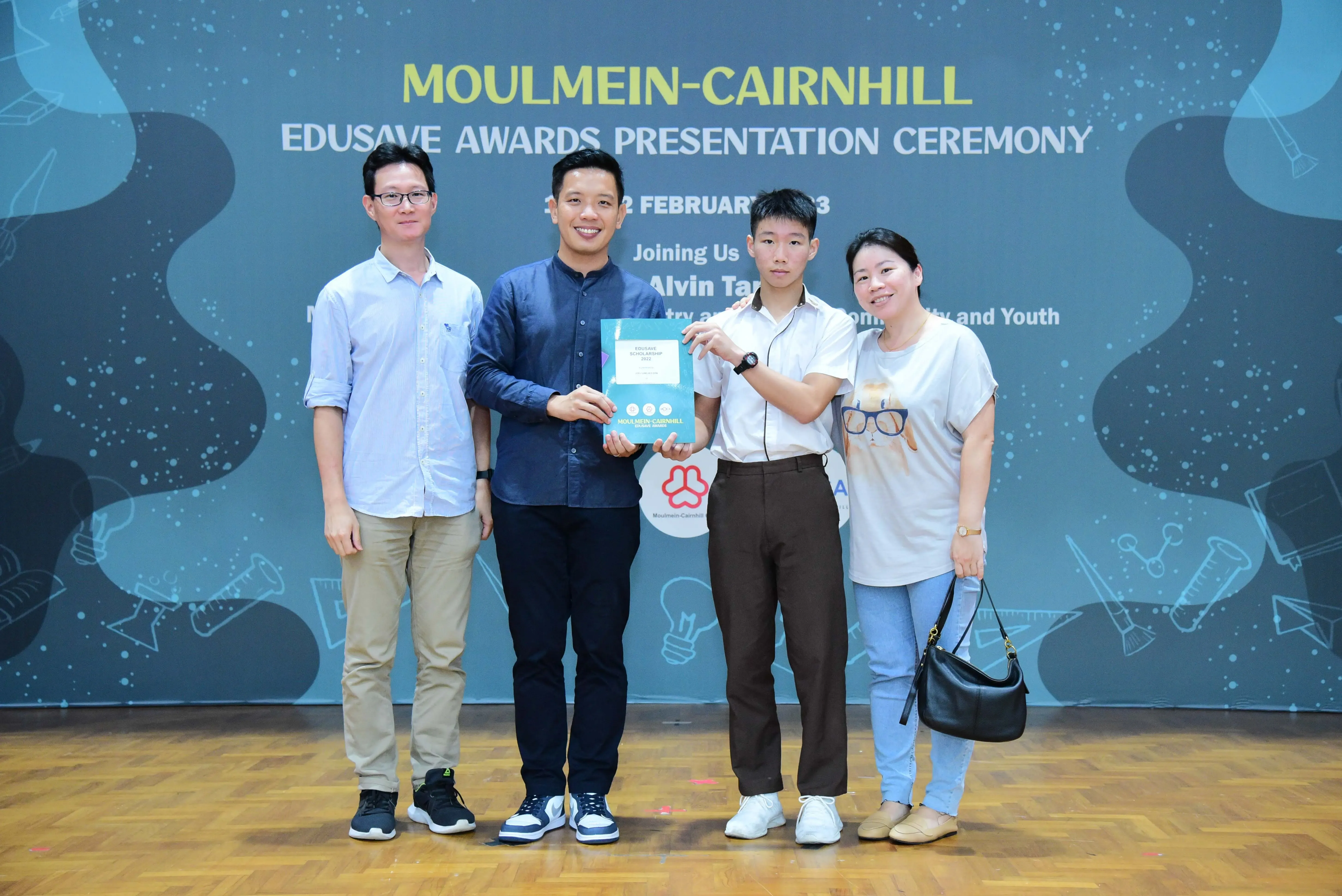 Four people pose holding a certificate at the Moulmein-Cairnhill Edusave Awards Presentation Ceremony backdrop.