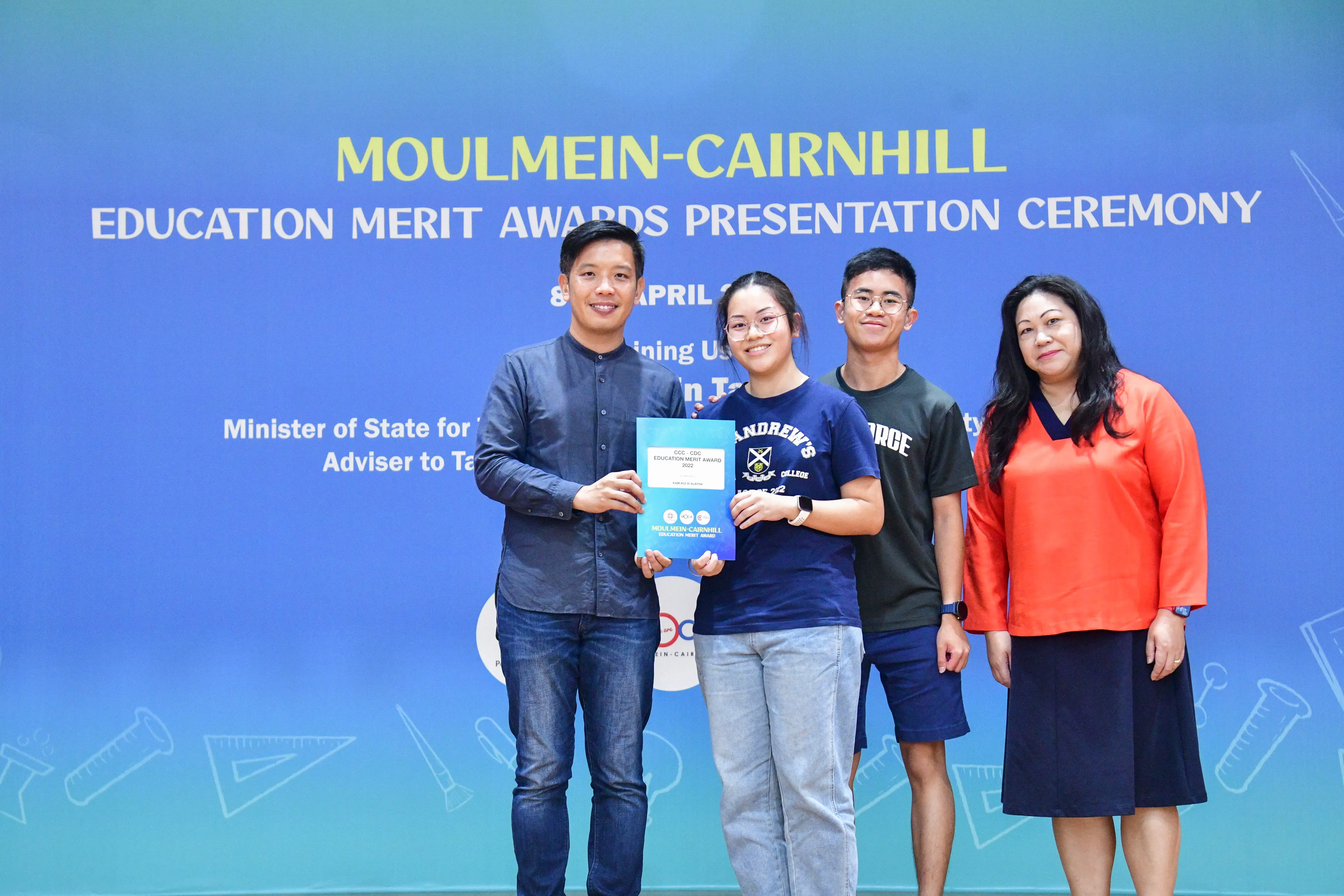 Four people pose in front of a blue Moulmein-Cairnhill Education Merit Awards Presentation Ceremony backdrop.