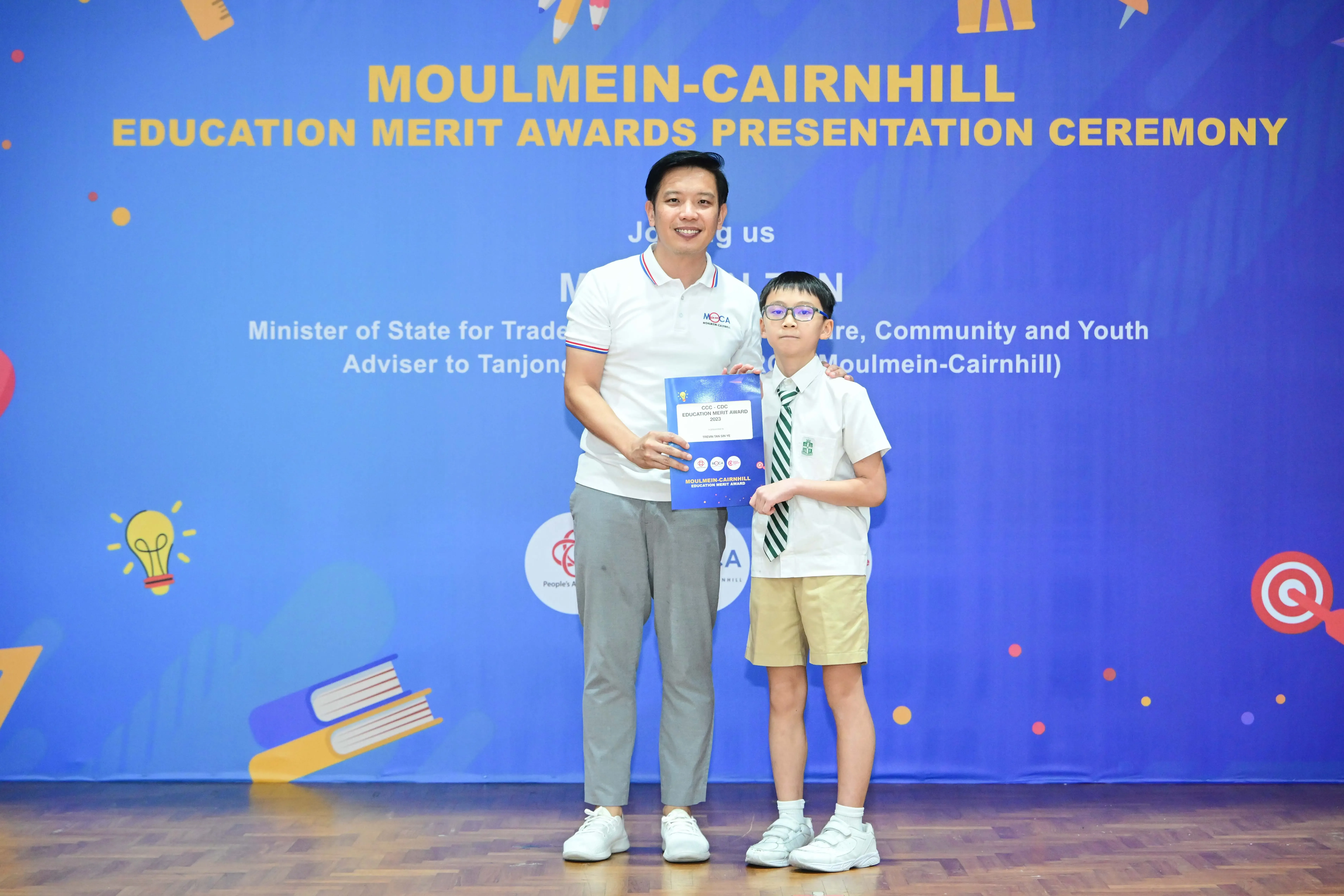 An adult and a young boy in school uniform hold an award certificate at the Moulmein-Cairnhill Education Merit Awards ceremony.
