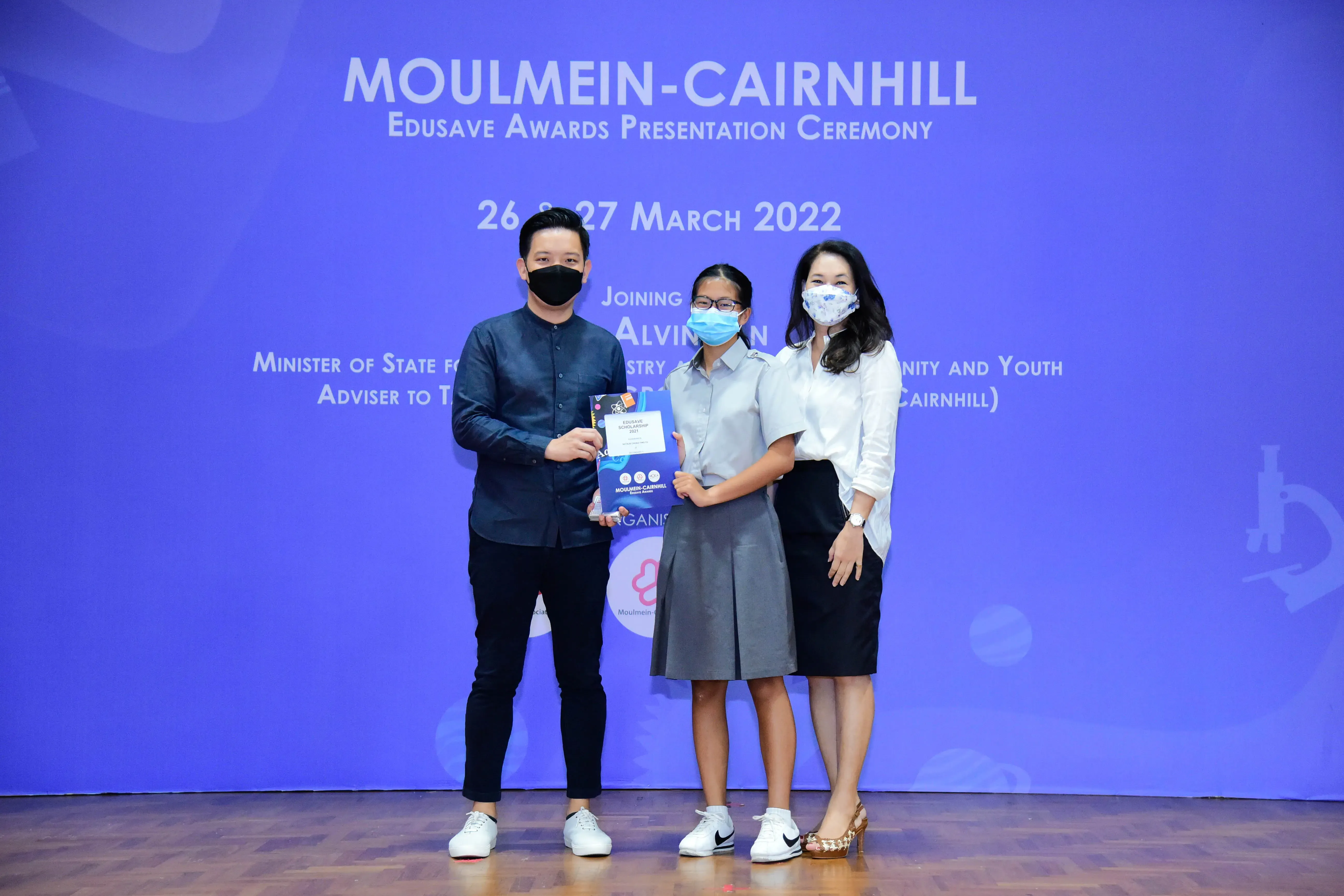 Three masked people pose on stage at the Moulmein-Cairnhill Edusave Awards Ceremony on 26-27 March 2022.