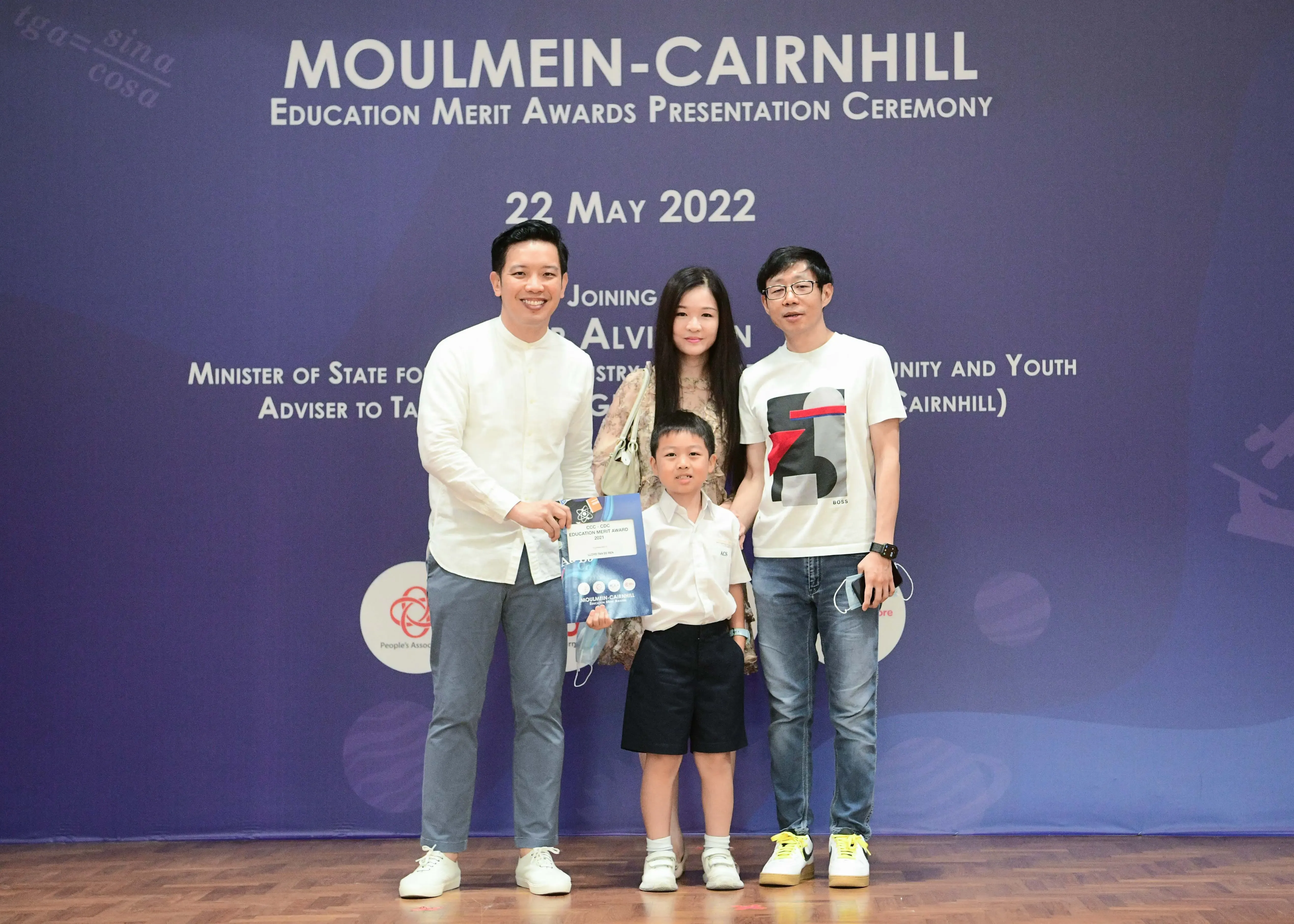 Three people pose before a Moulmein-Cairnhill Education Merit Awards banner dated 22 May 2022, with a child holding an award.