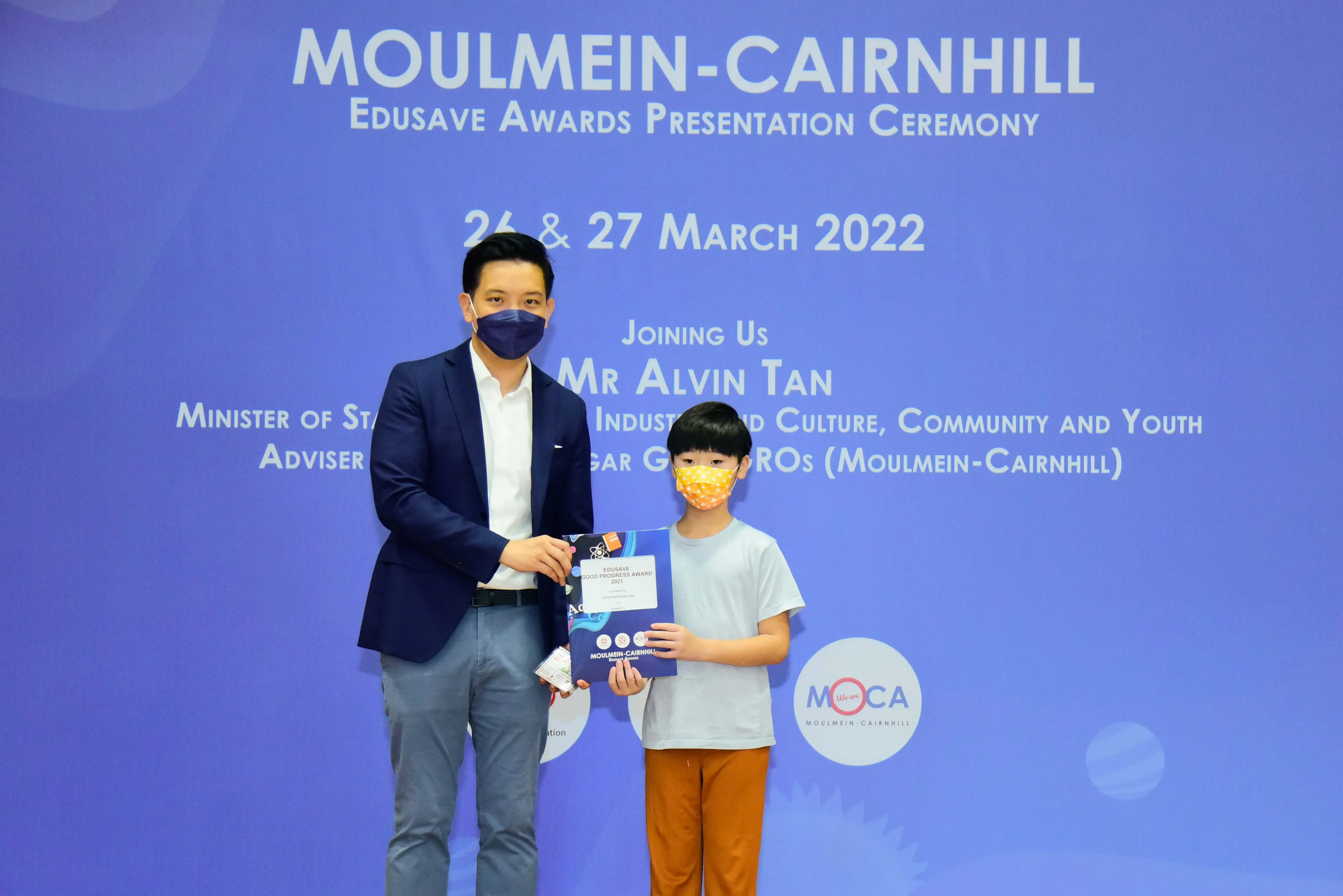 An adult and a child wearing masks hold an award certificate at the Moulmein-Cairnhill Edusave Awards Ceremony 2022.
