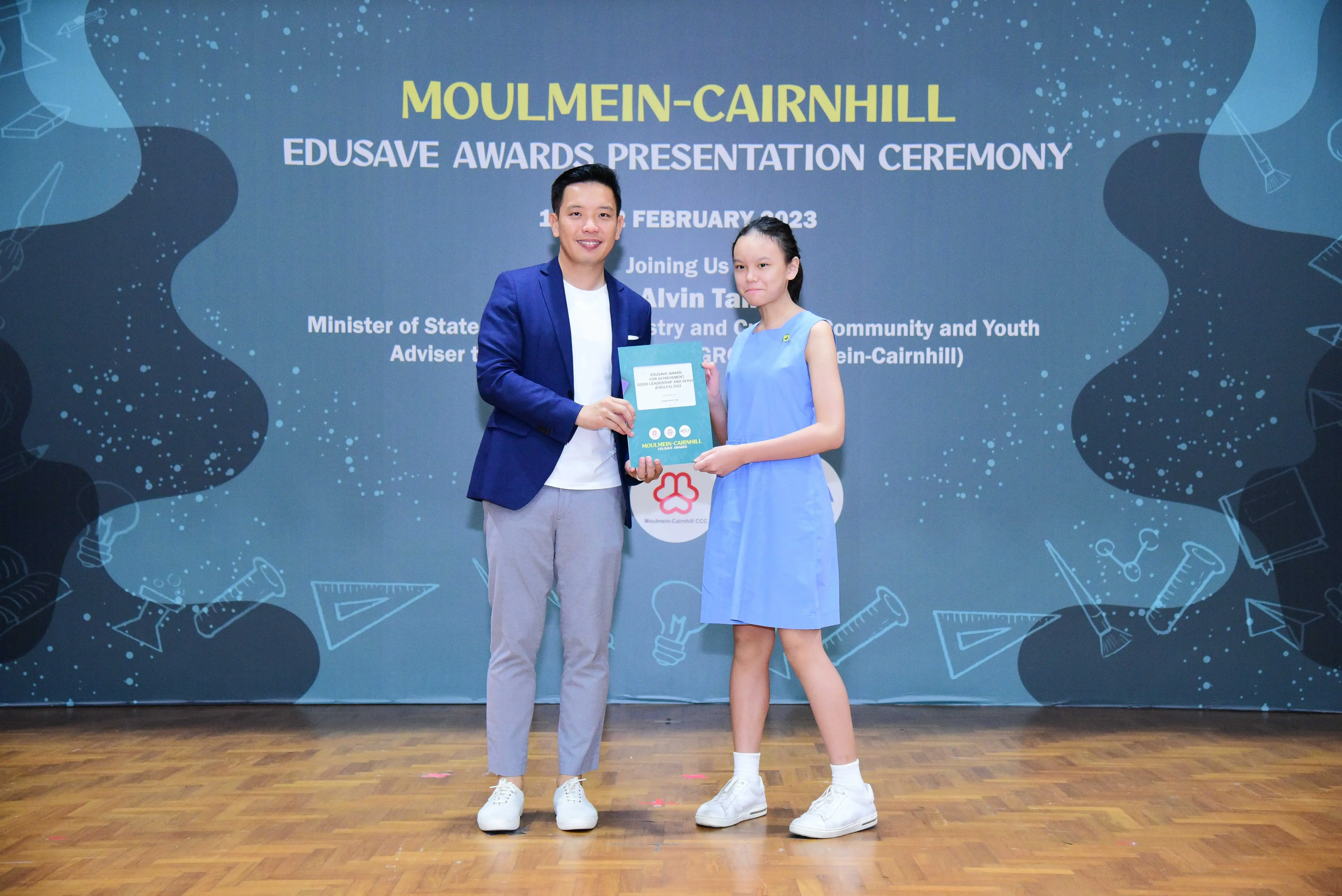 A man in a blue blazer presents an award to a girl in a blue dress at the Moulmein-Cairnhill Edusave Awards Ceremony 2023.