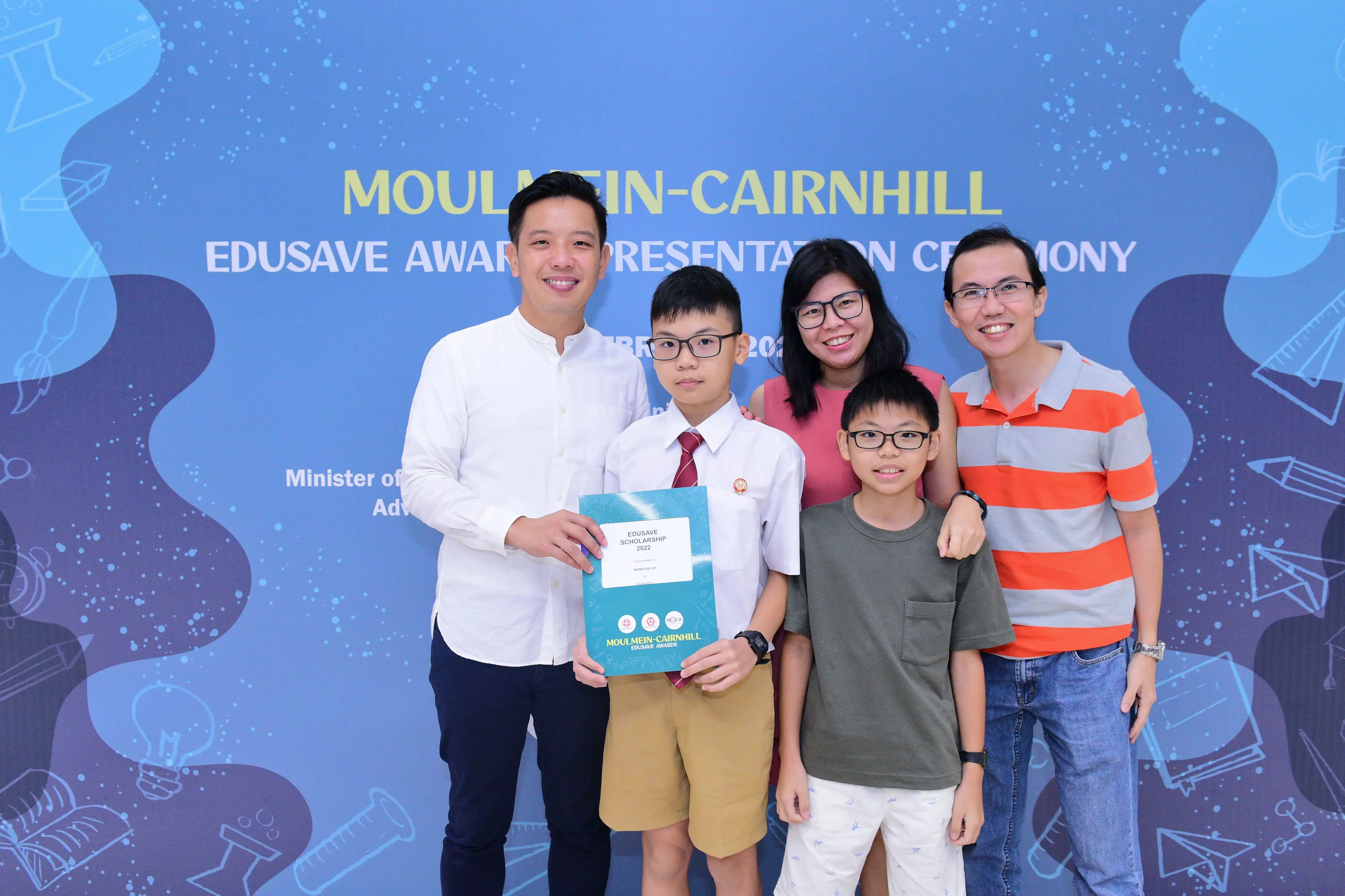 Five people pose smiling at the Moulmein-Cairnhill Edusave Awards Presentation Ceremony, with a boy holding an award folder.