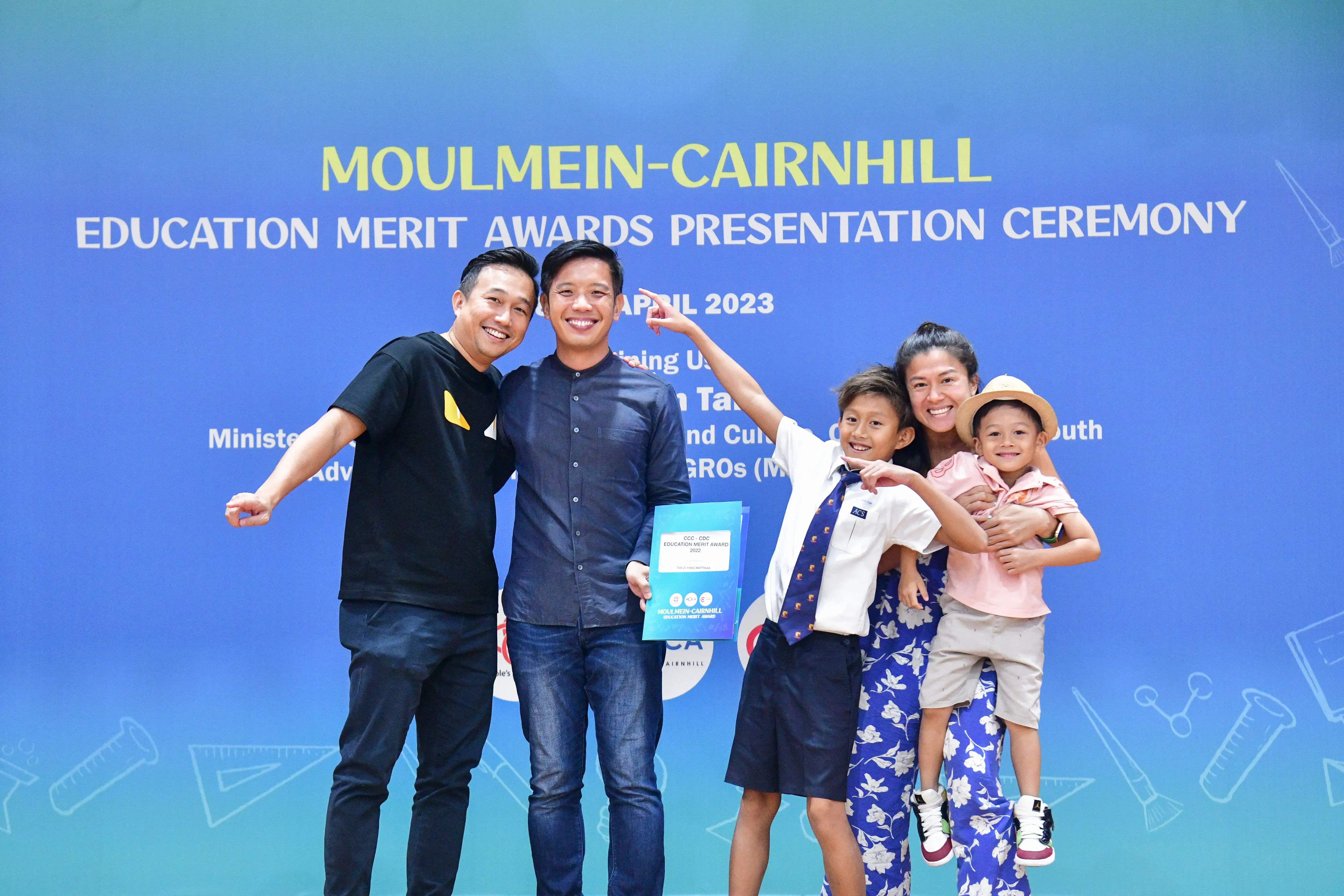 Five smiling people pose before a Moulmein-Cairnhill Education Merit Awards Presentation Ceremony banner, April 2023.