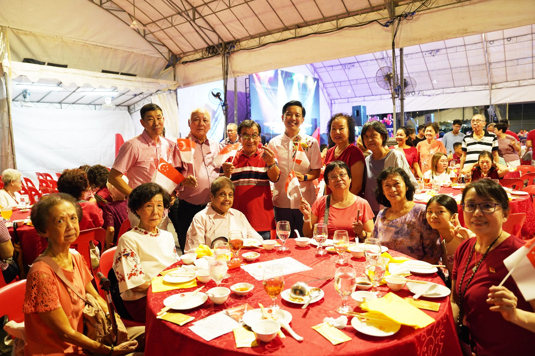 A group of people seated and standing around a red-clothed dining table, waving small Singapore flags at an outdoor event.