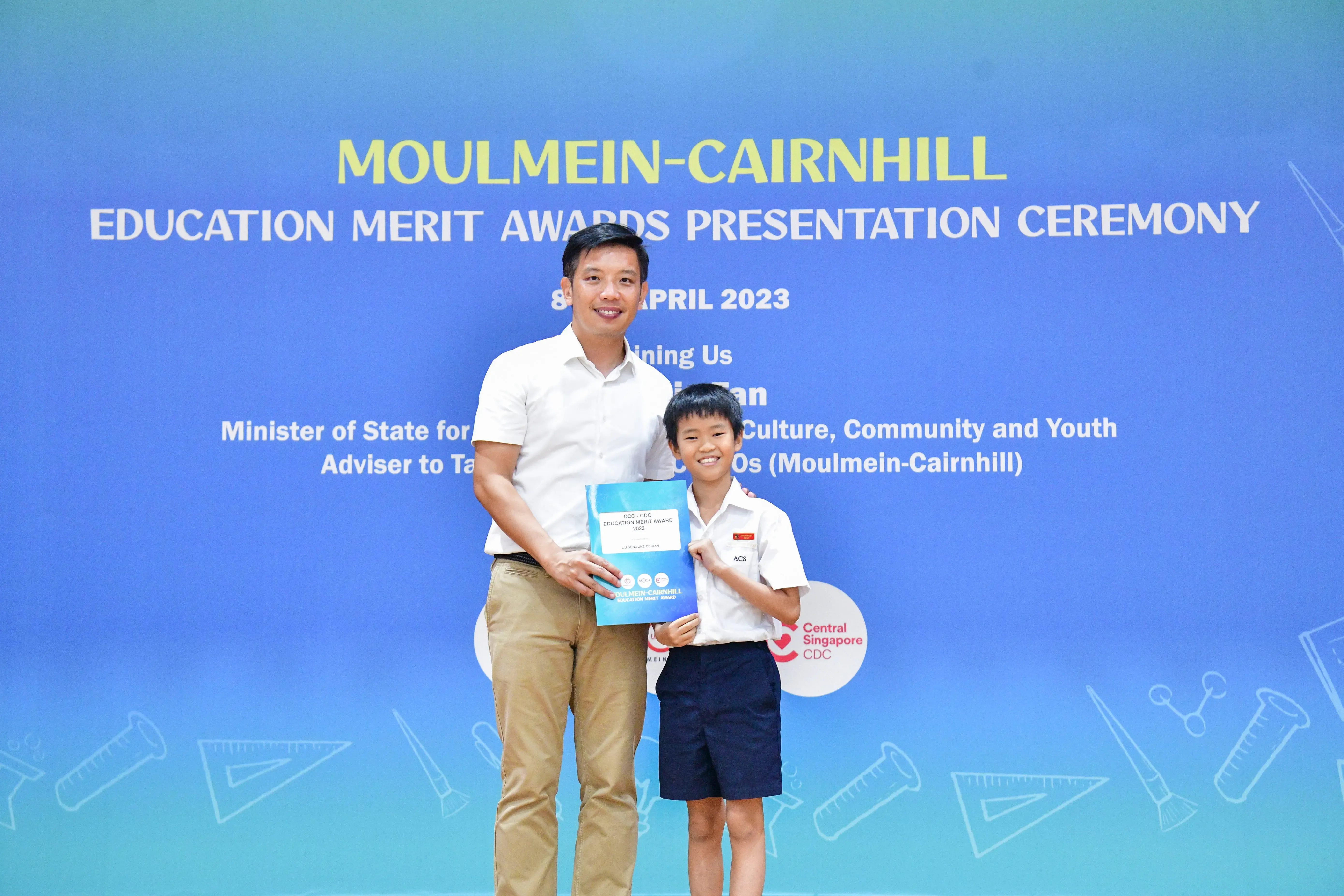 An adult man and a young boy hold a certificate together at the Moulmein-Cairnhill Education Merit Awards Ceremony 2023.