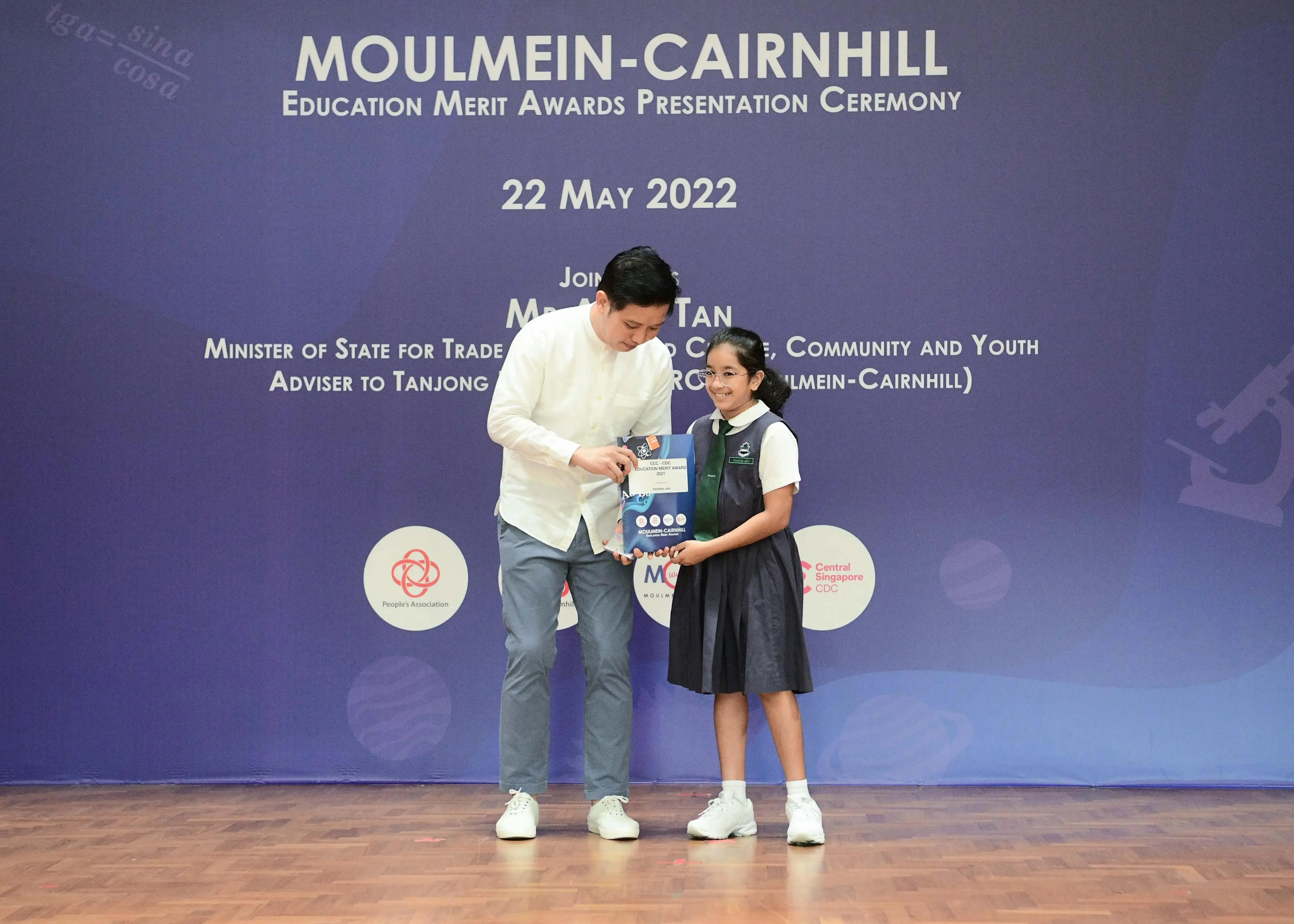 A man in white shirt presents an award to a young girl in school uniform at Moulmein-Cairnhill Education Merit Awards 2022.