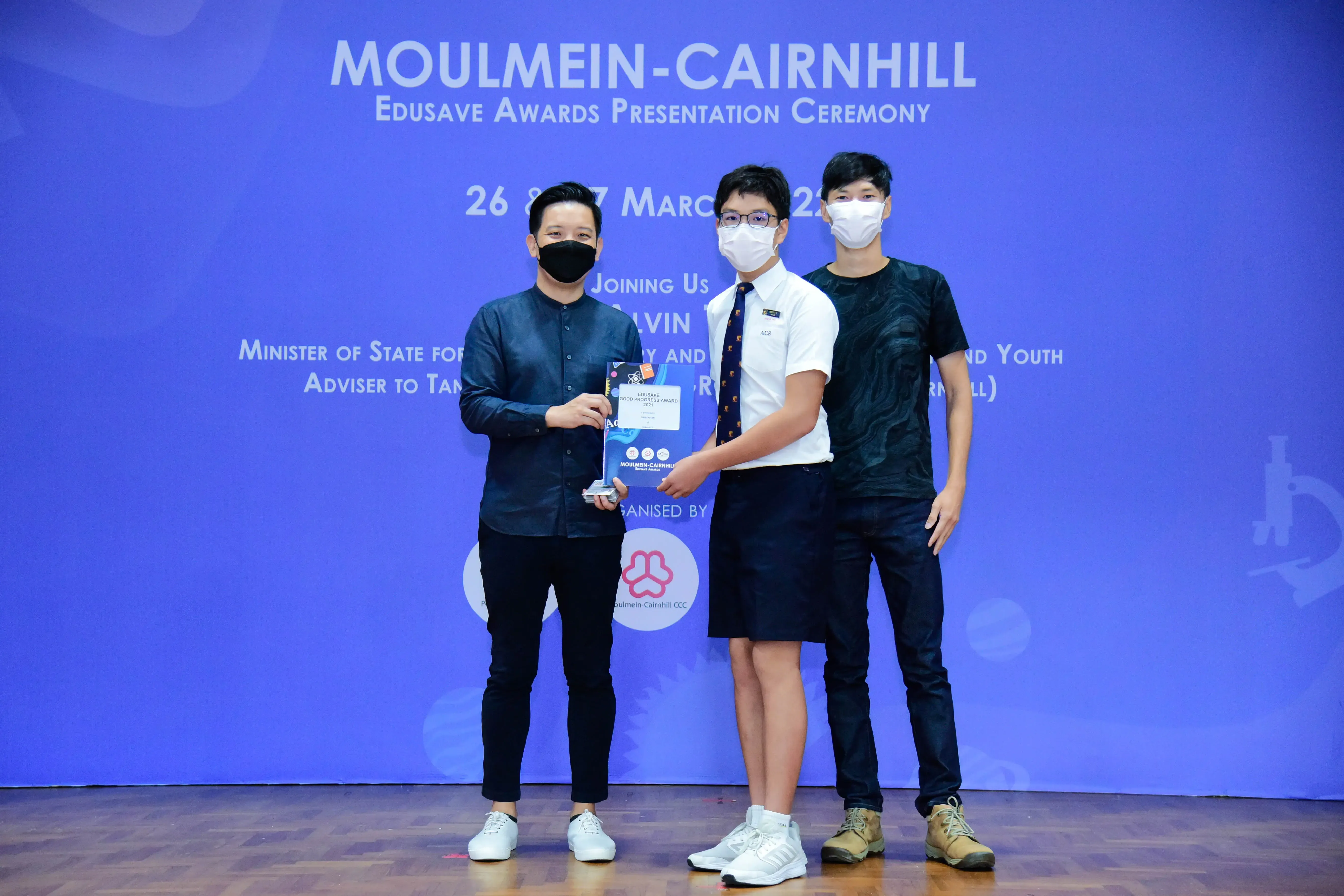 Three masked people stand on a stage at the Moulmein-Cairnhill Edusave Awards Presentation Ceremony, holding a trophy.