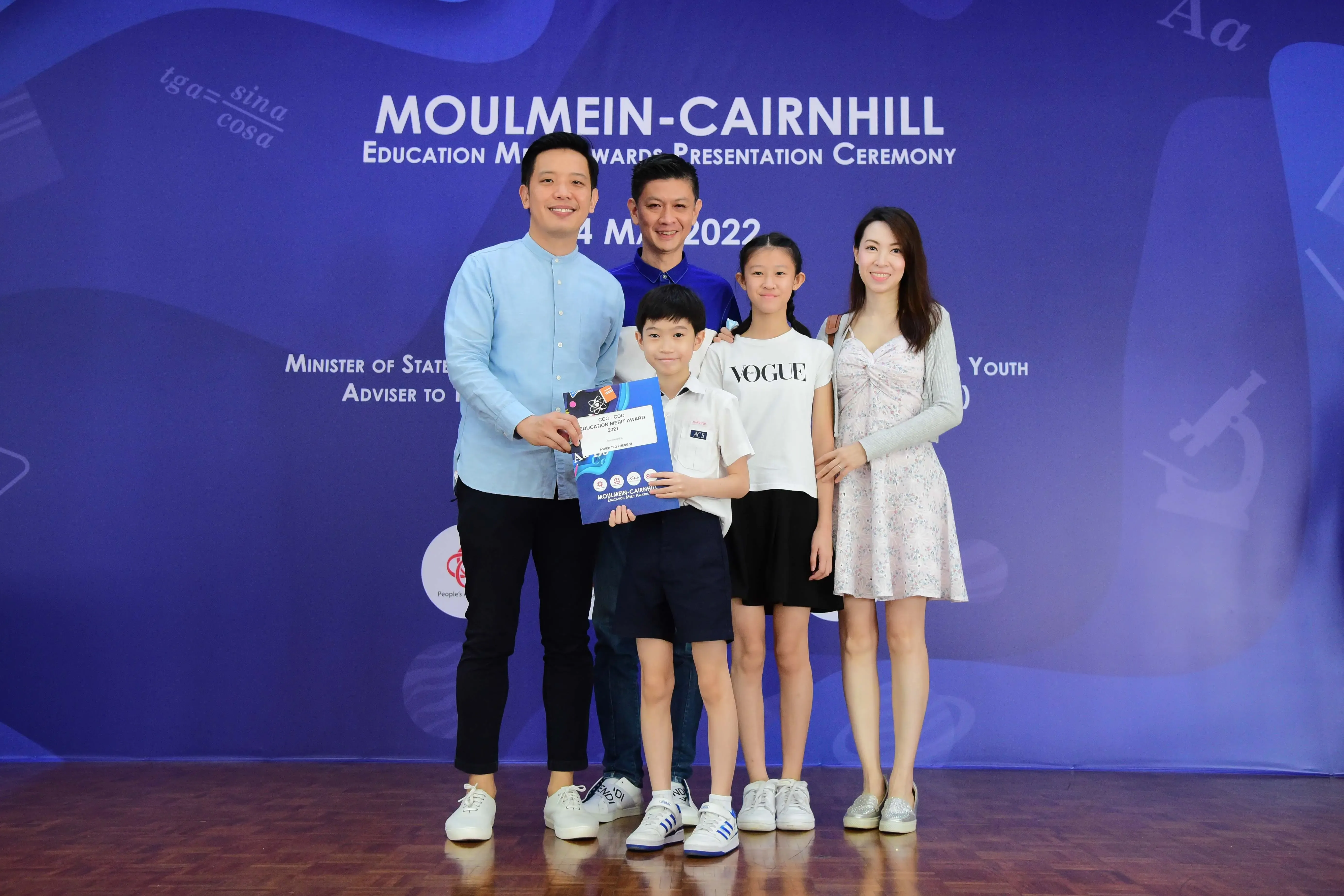Five people pose in front of a Moulmein-Cairnhill Education Merit Awards Presentation Ceremony backdrop dated 2022.