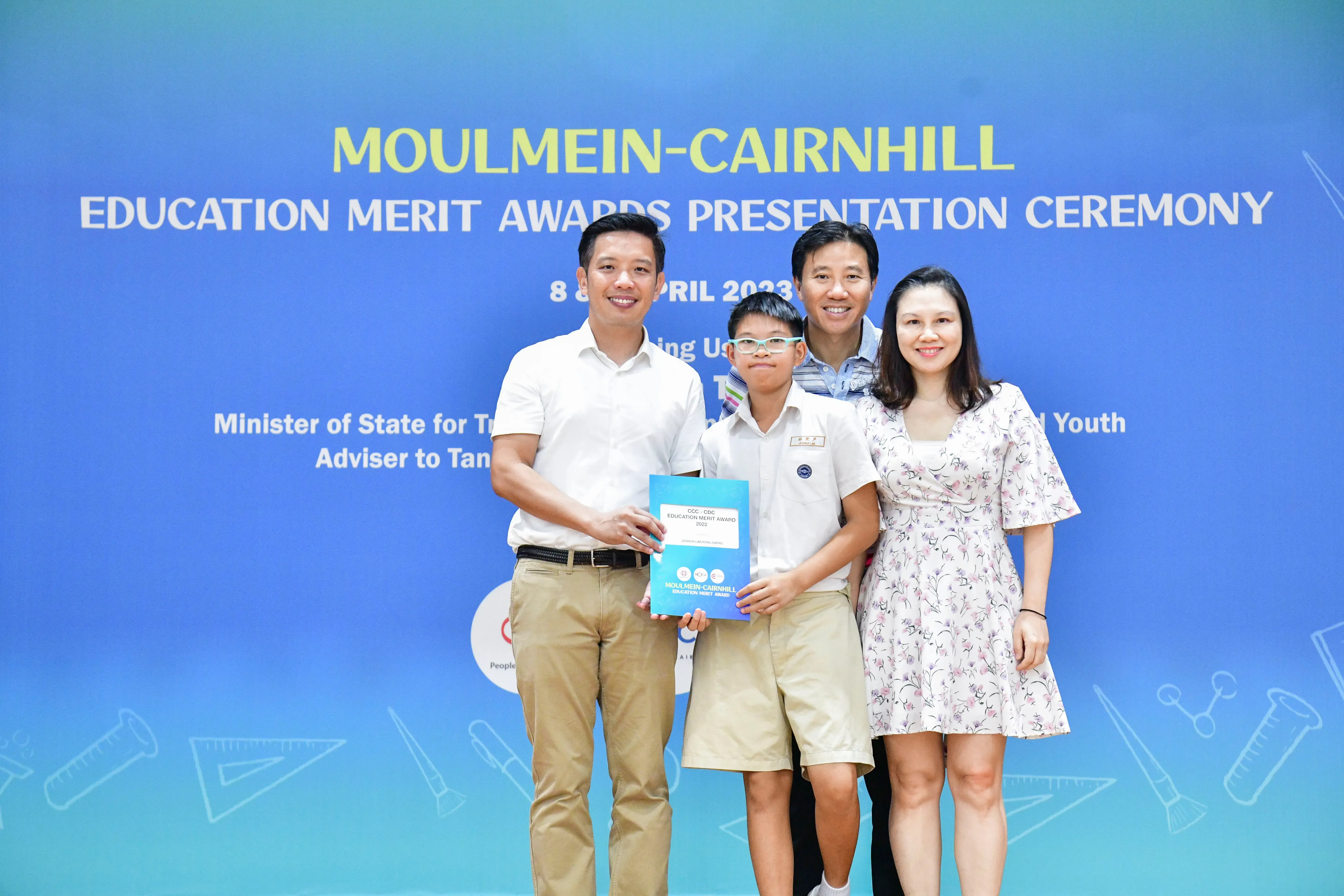 Four people pose at the Moulmein-Cairnhill Education Merit Awards Presentation Ceremony, with a boy holding a blue award folder.