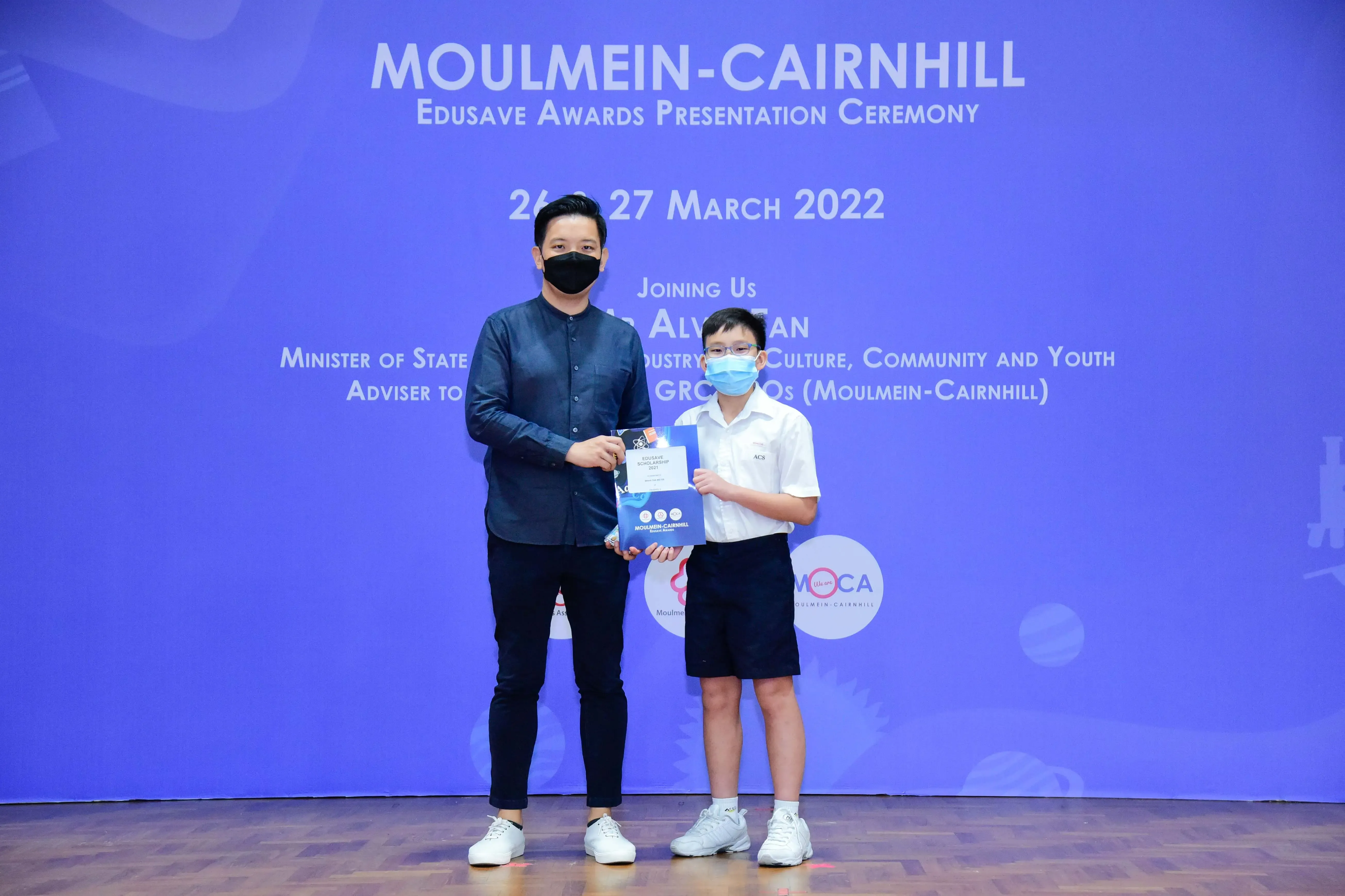 An adult and a young student wearing masks hold a certificate at the Moulmein-Cairnhill Edusave Awards Ceremony 2022.
