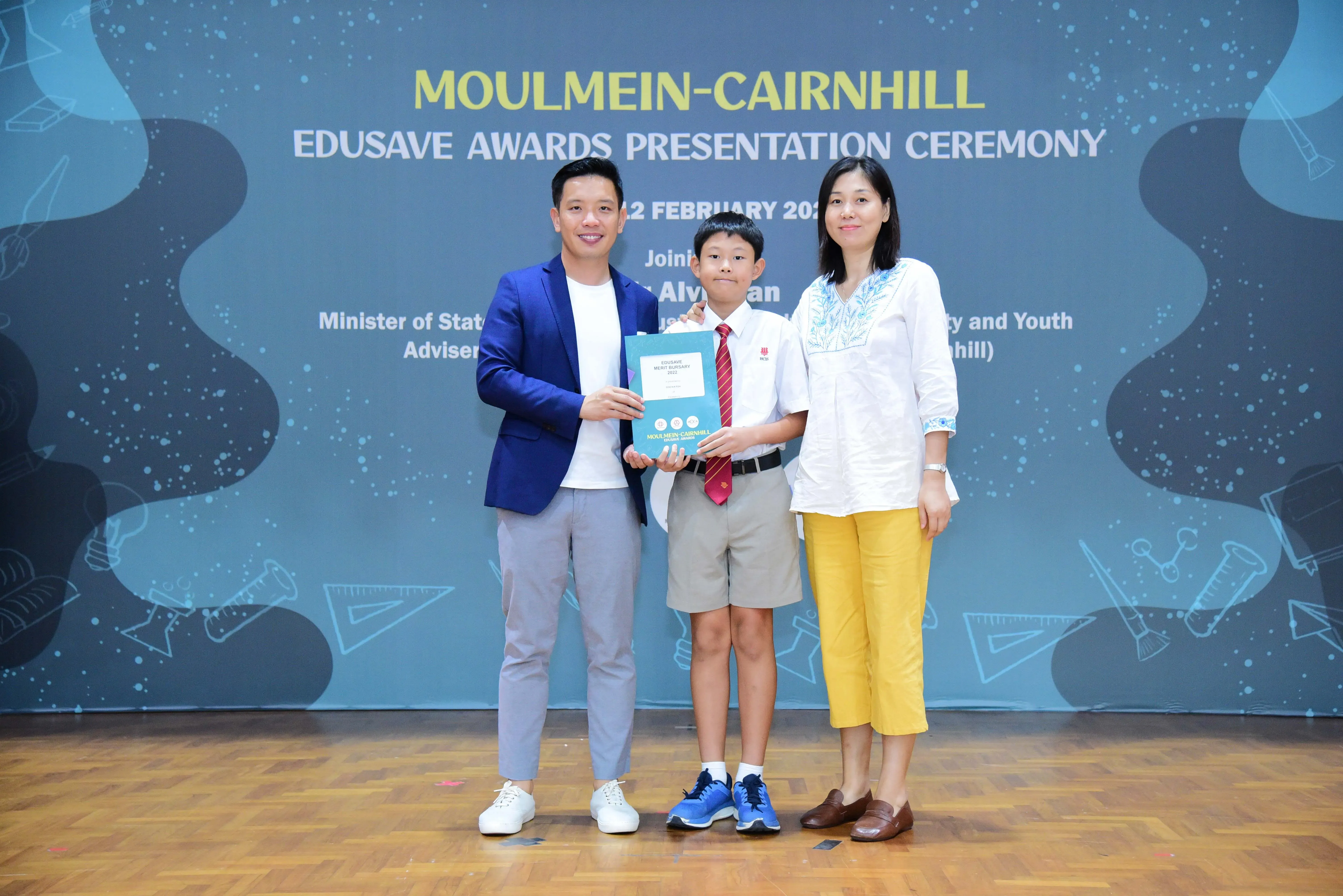 A boy in school uniform holds an award booklet flanked by two adults at the Moulmein-Cairnhill Edusave Awards Ceremony.