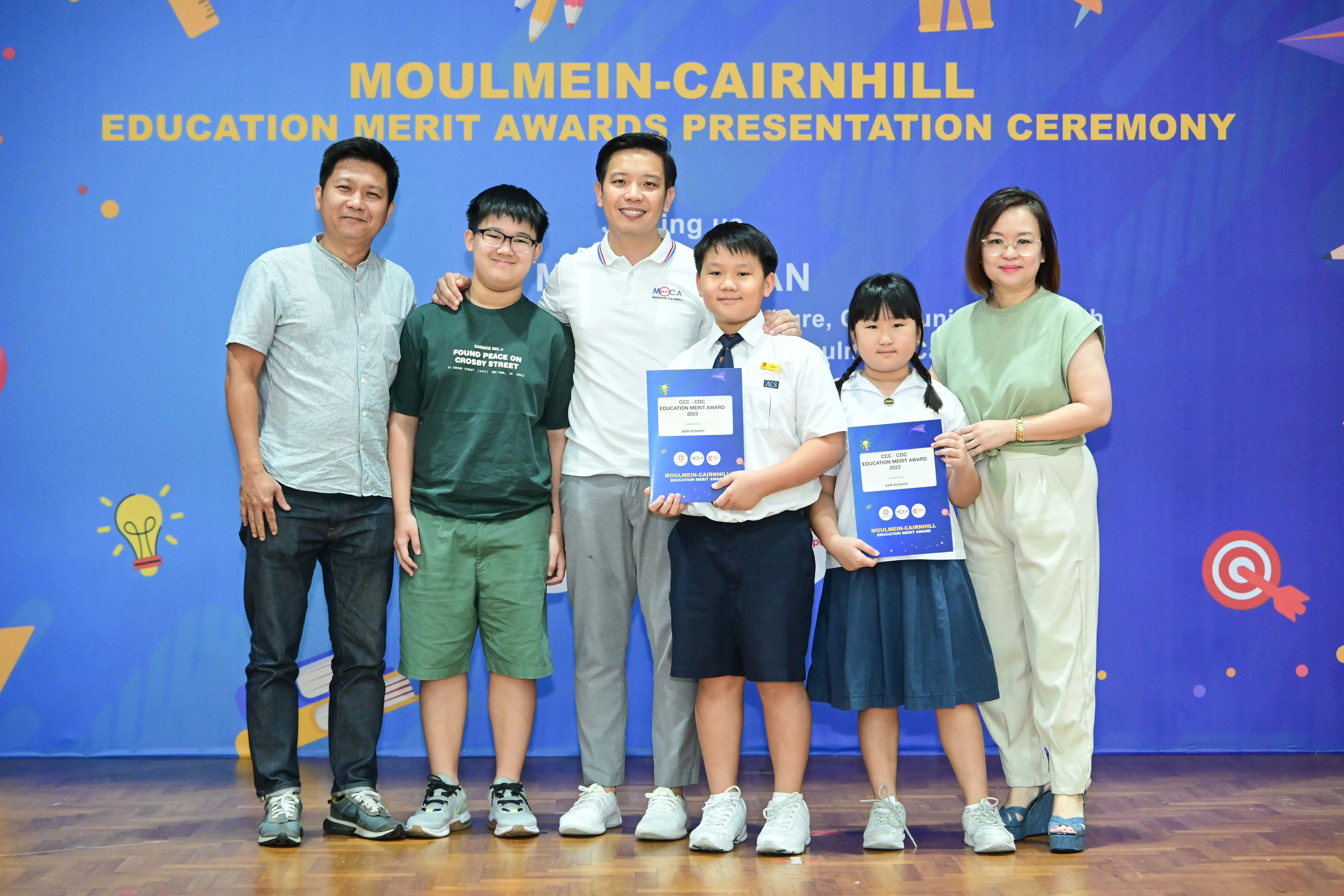 Six people pose at the Moulmein-Cairnhill Education Merit Awards Presentation Ceremony, two children holding award certificates.