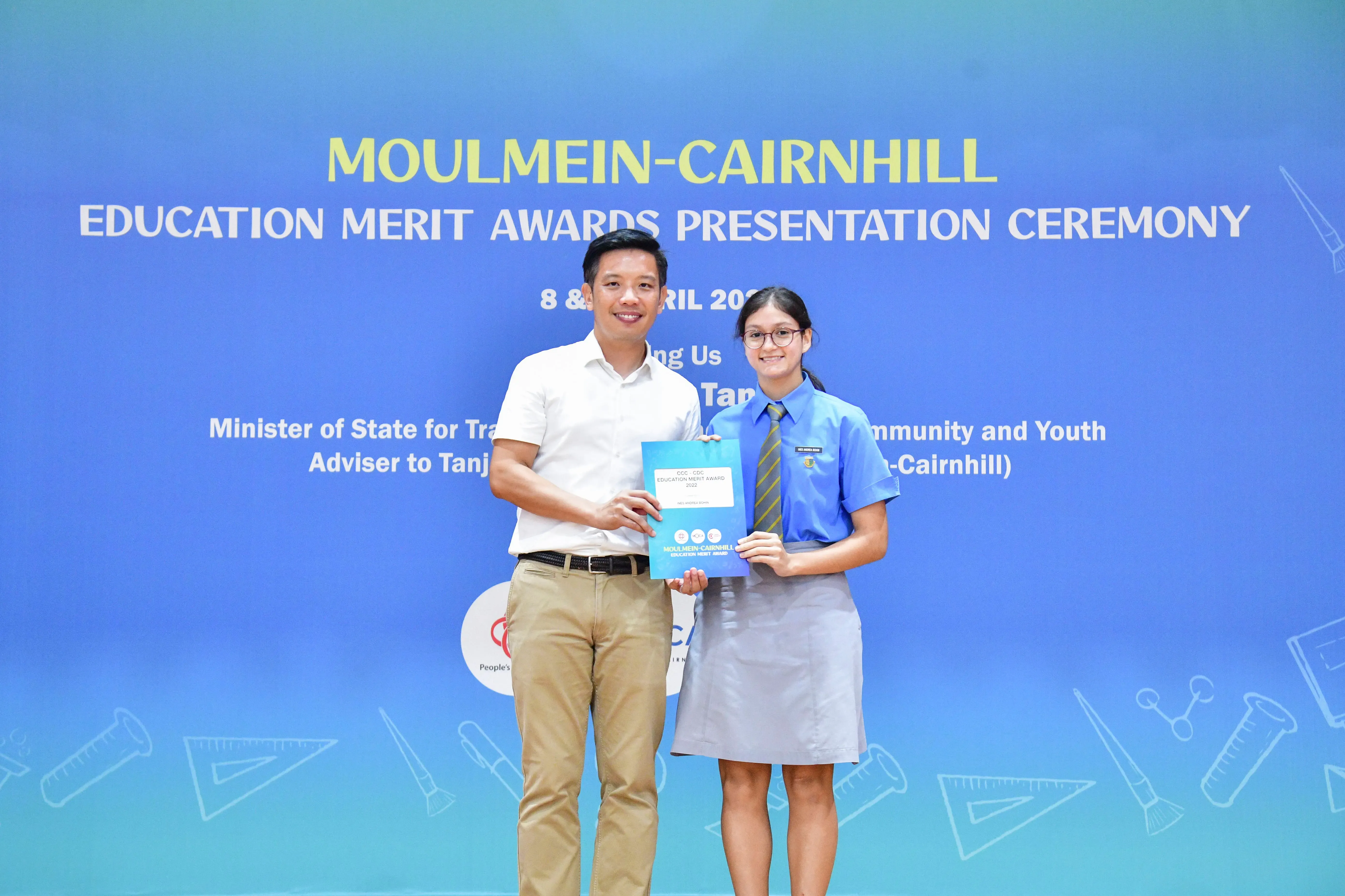 Two people hold a blue award folder in front of a Moulmein-Cairnhill Education Merit Awards Presentation Ceremony banner.