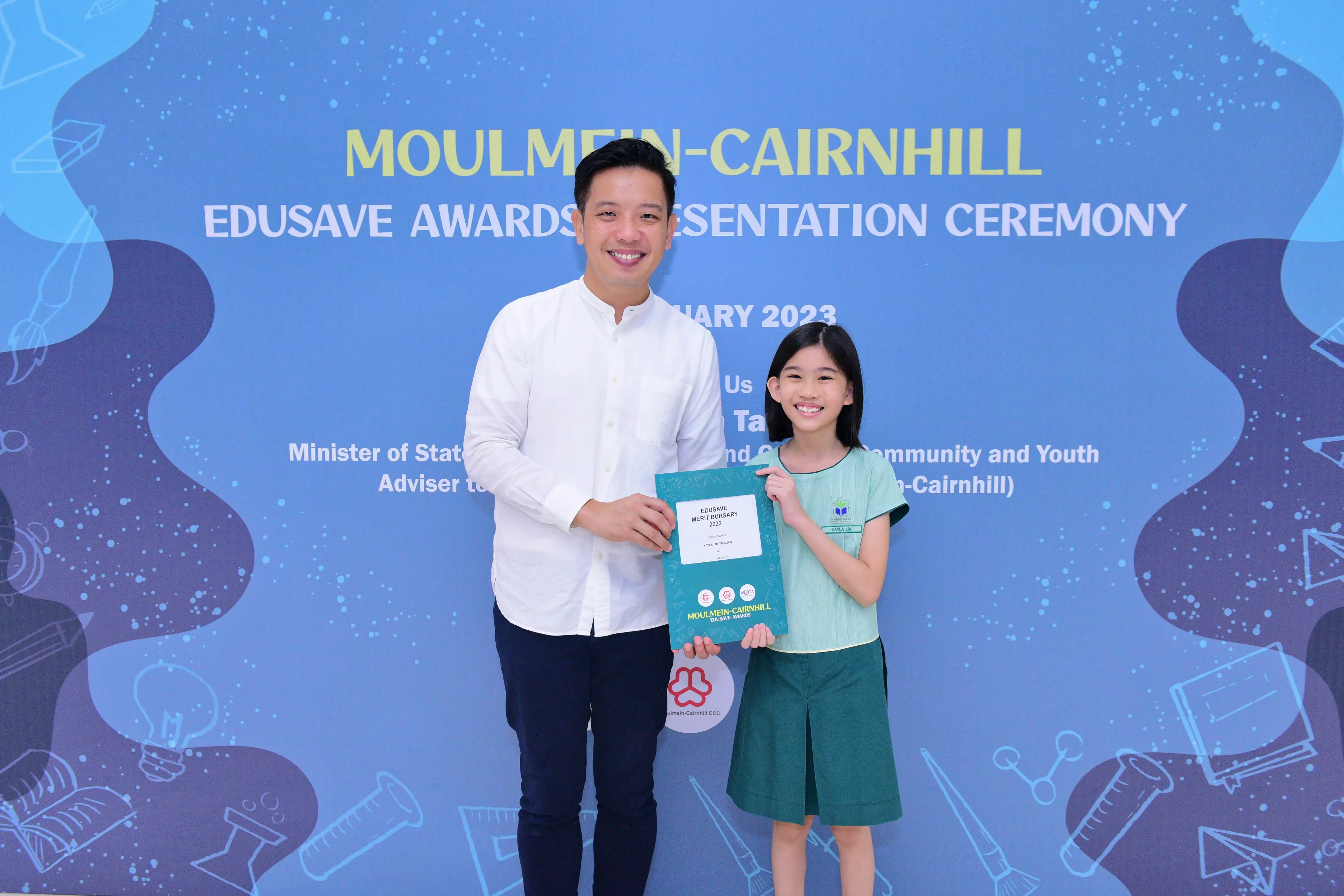A man and a young girl hold a teal award folder at the Moulmein-Cairnhill Edusave Awards Presentation Ceremony 2023.