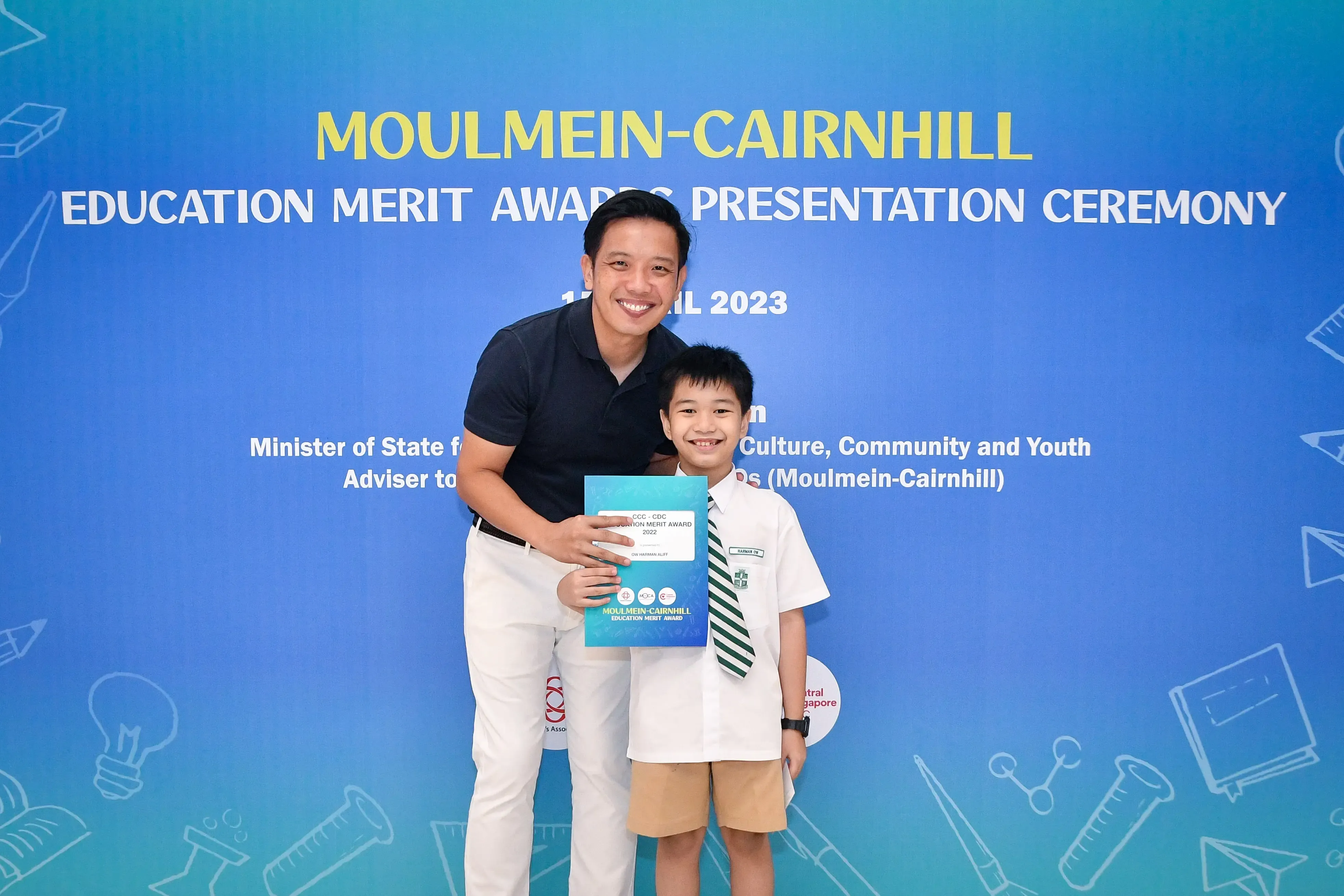 A man and a young boy in school uniform hold an award certificate at the 2023 Moulmein-Cairnhill Education Merit Awards ceremony.