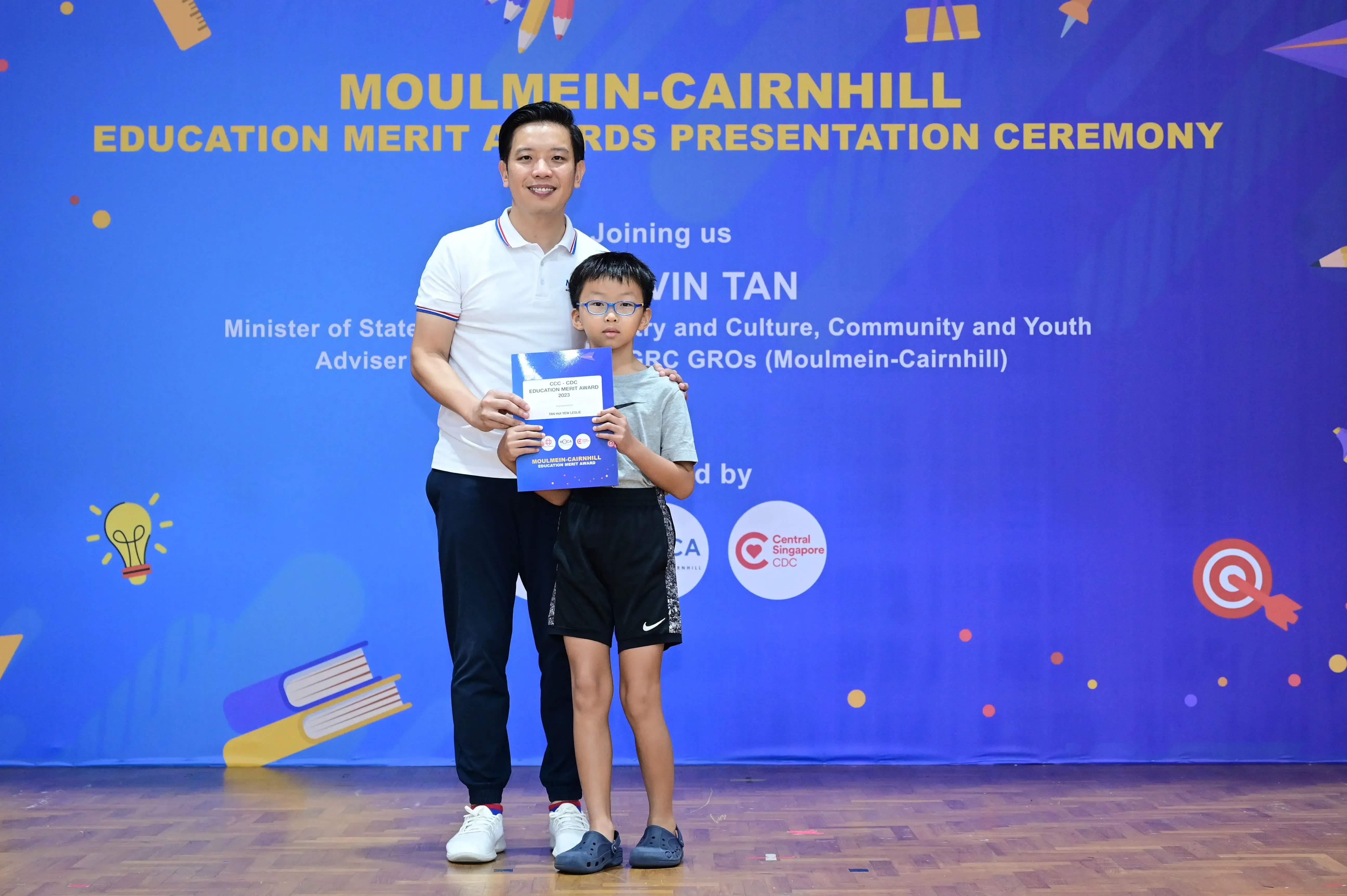 A man and a young boy holding an award certificate stand before a Moulmein-Cairnhill Education Merit Awards banner.