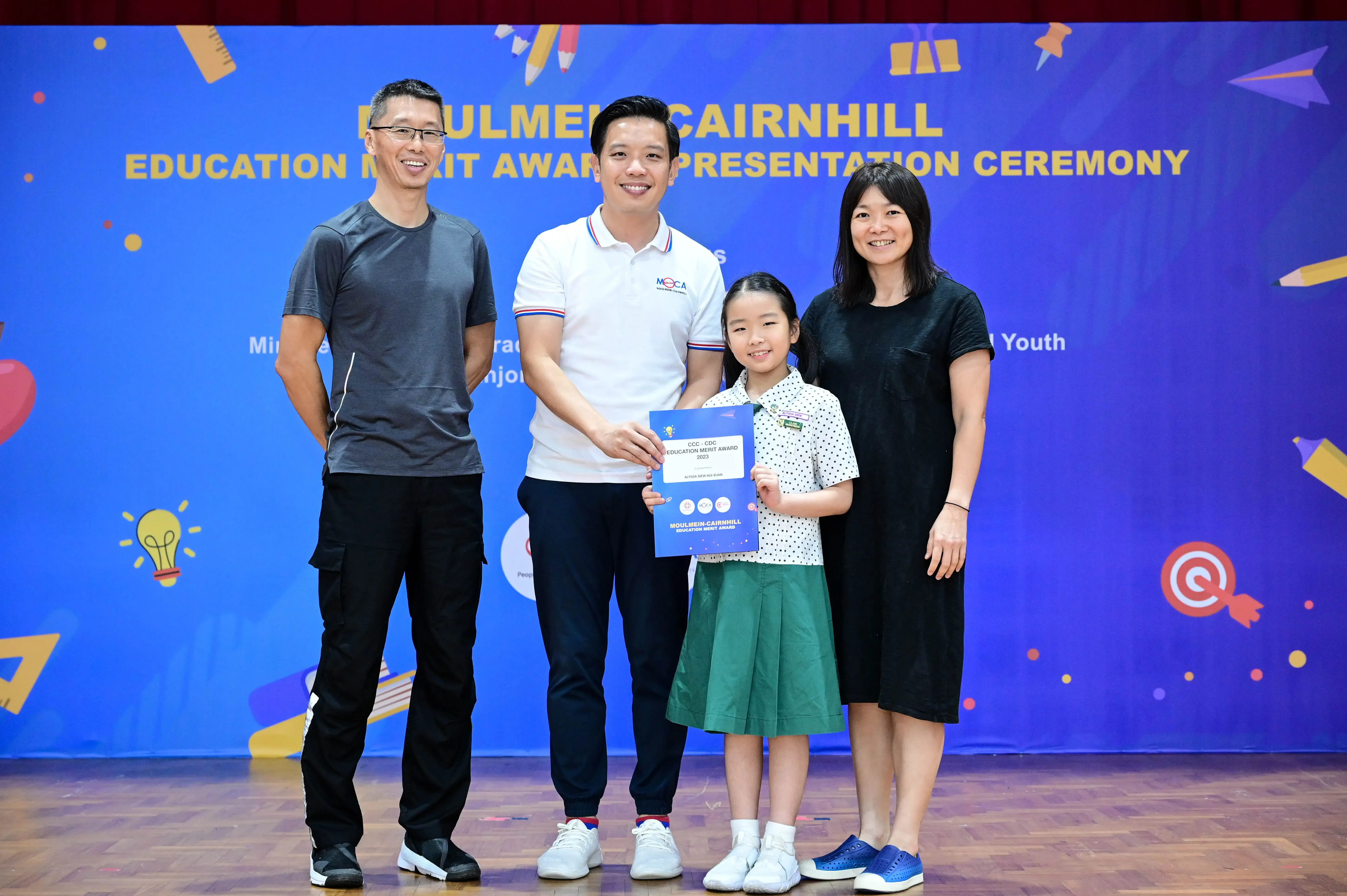 A young girl holds a certificate flanked by three adults at the Moulmein-Cairnhill Education Merit Award Ceremony.