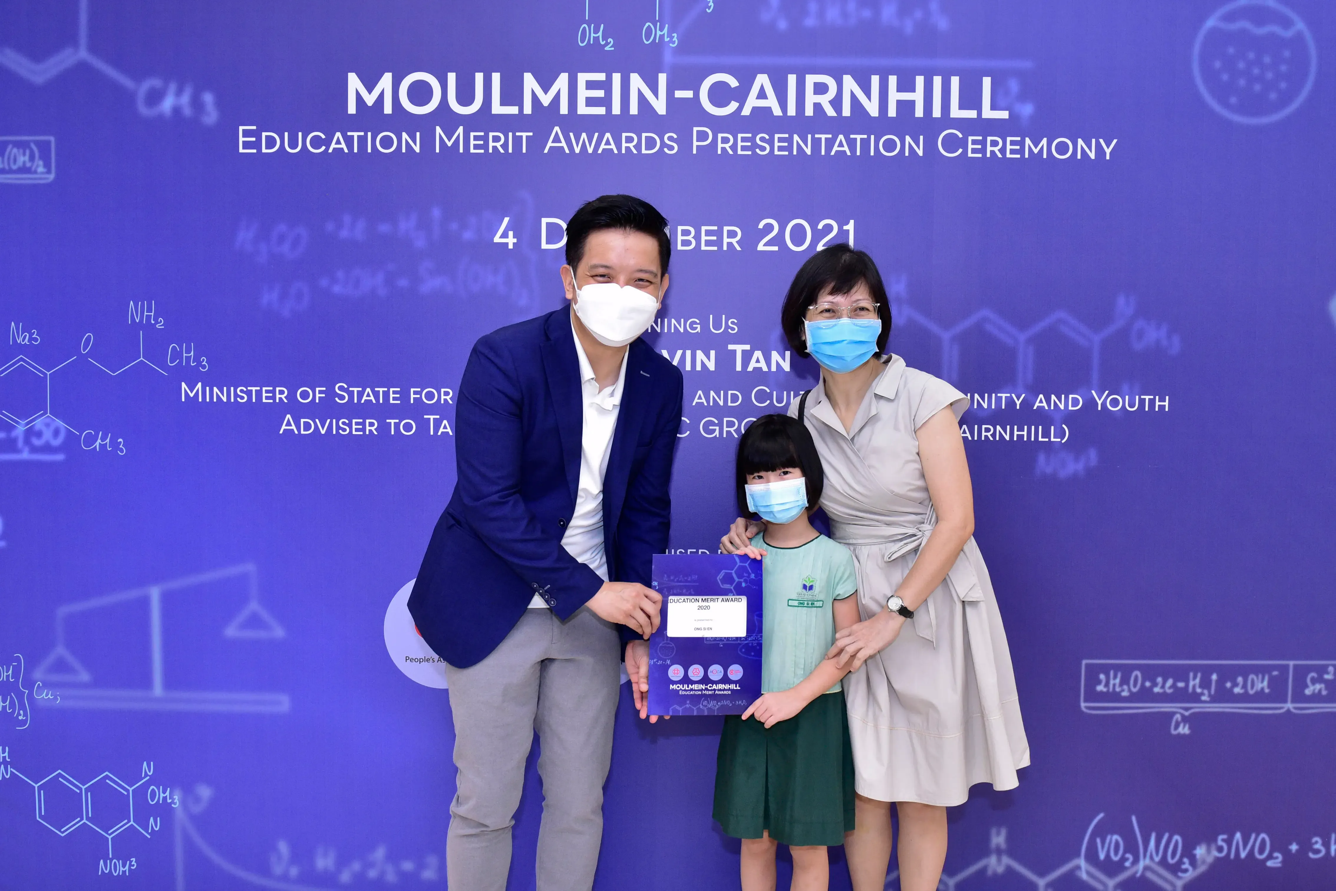 Two masked adults and a masked child hold an award certificate at the Moulmein-Cairnhill Education Merit Awards 2021.