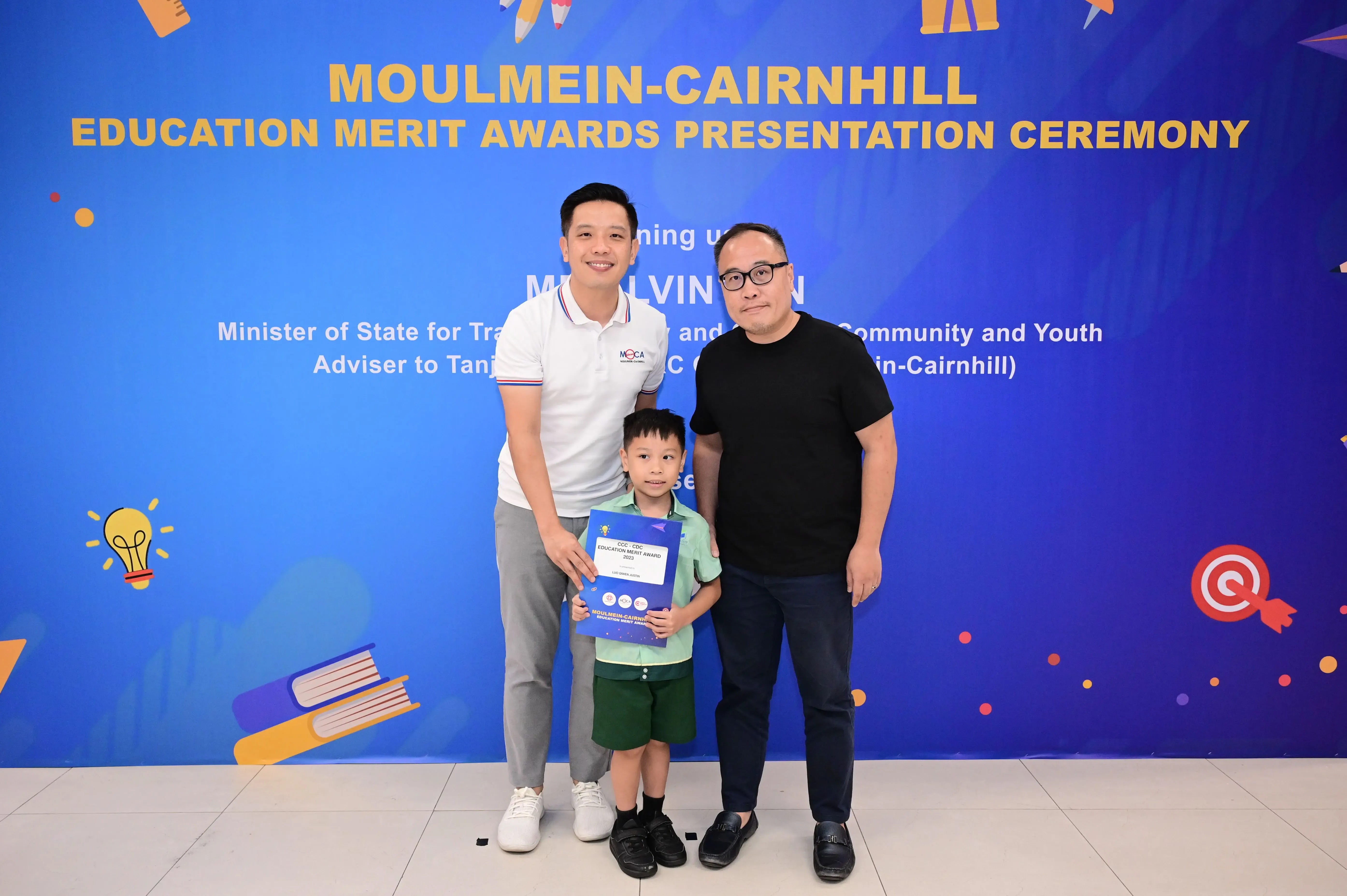 Two men and a young boy holding an award certificate pose before a Moulmein-Cairnhill Education Merit Awards banner.