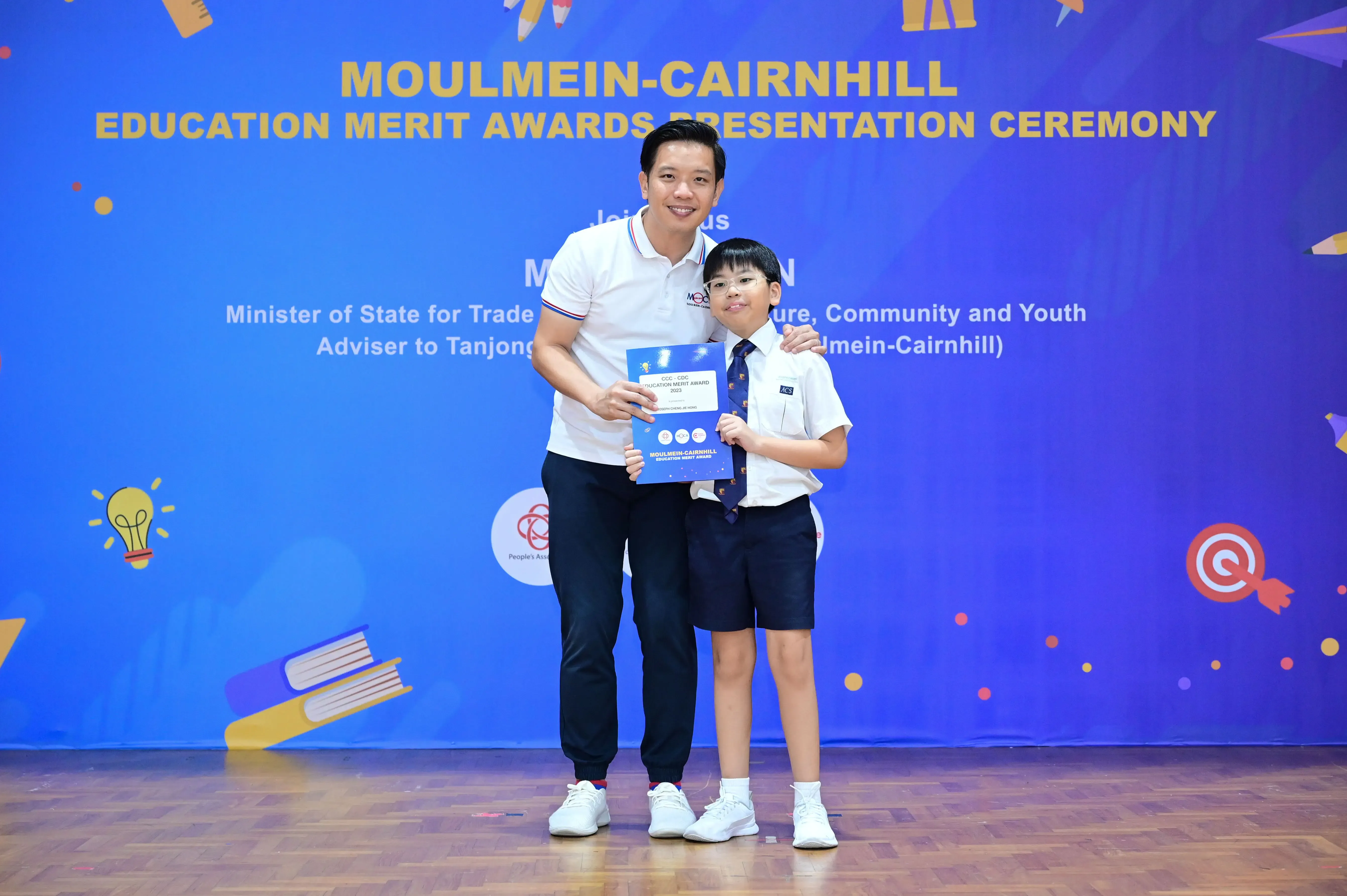 An adult and a young boy in school uniform hold a certificate at the Moulmein-Cairnhill Education Merit Awards ceremony.