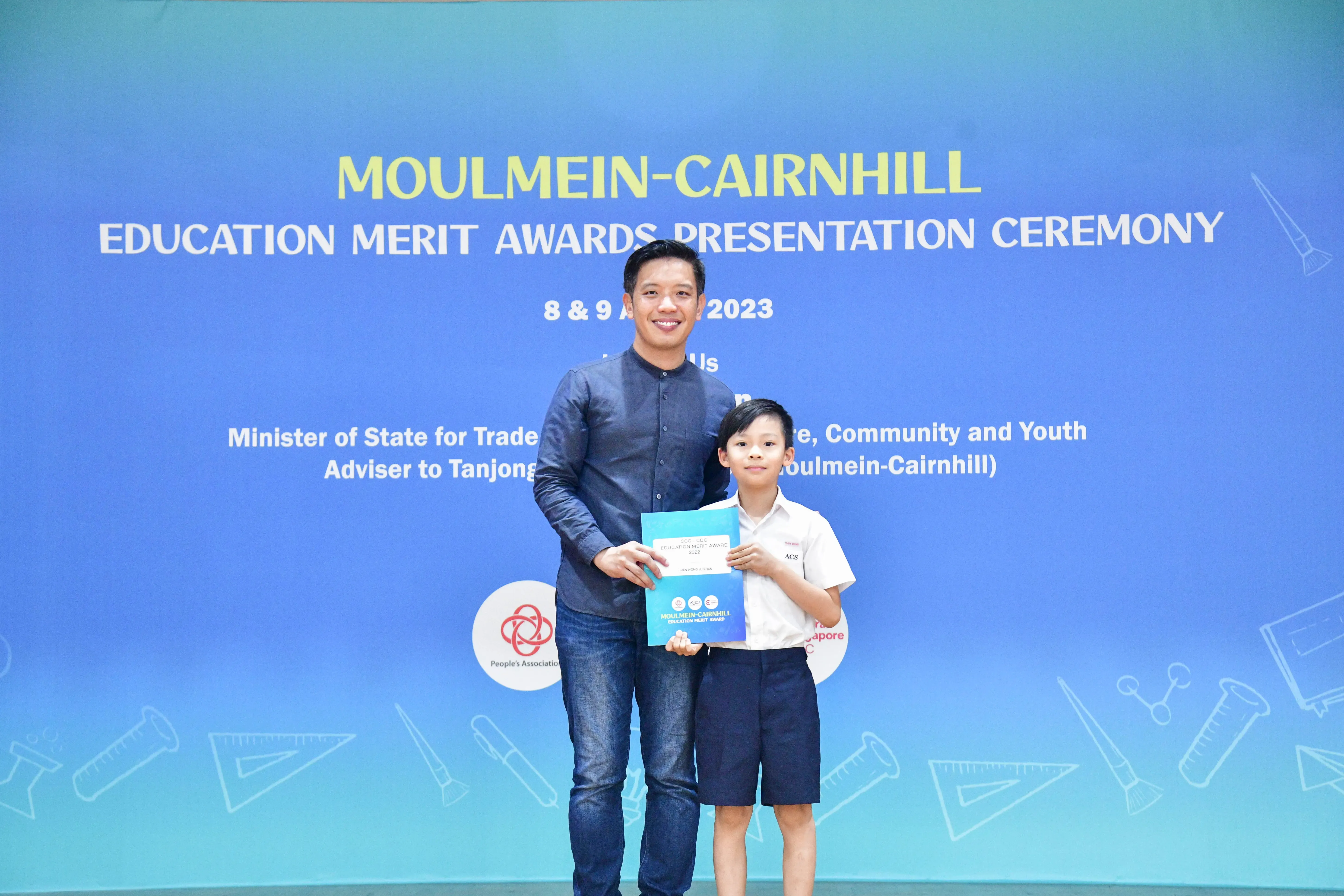 A man and a young boy hold an award certificate in front of a Moulmein-Cairnhill Education Merit Awards 2023 banner.