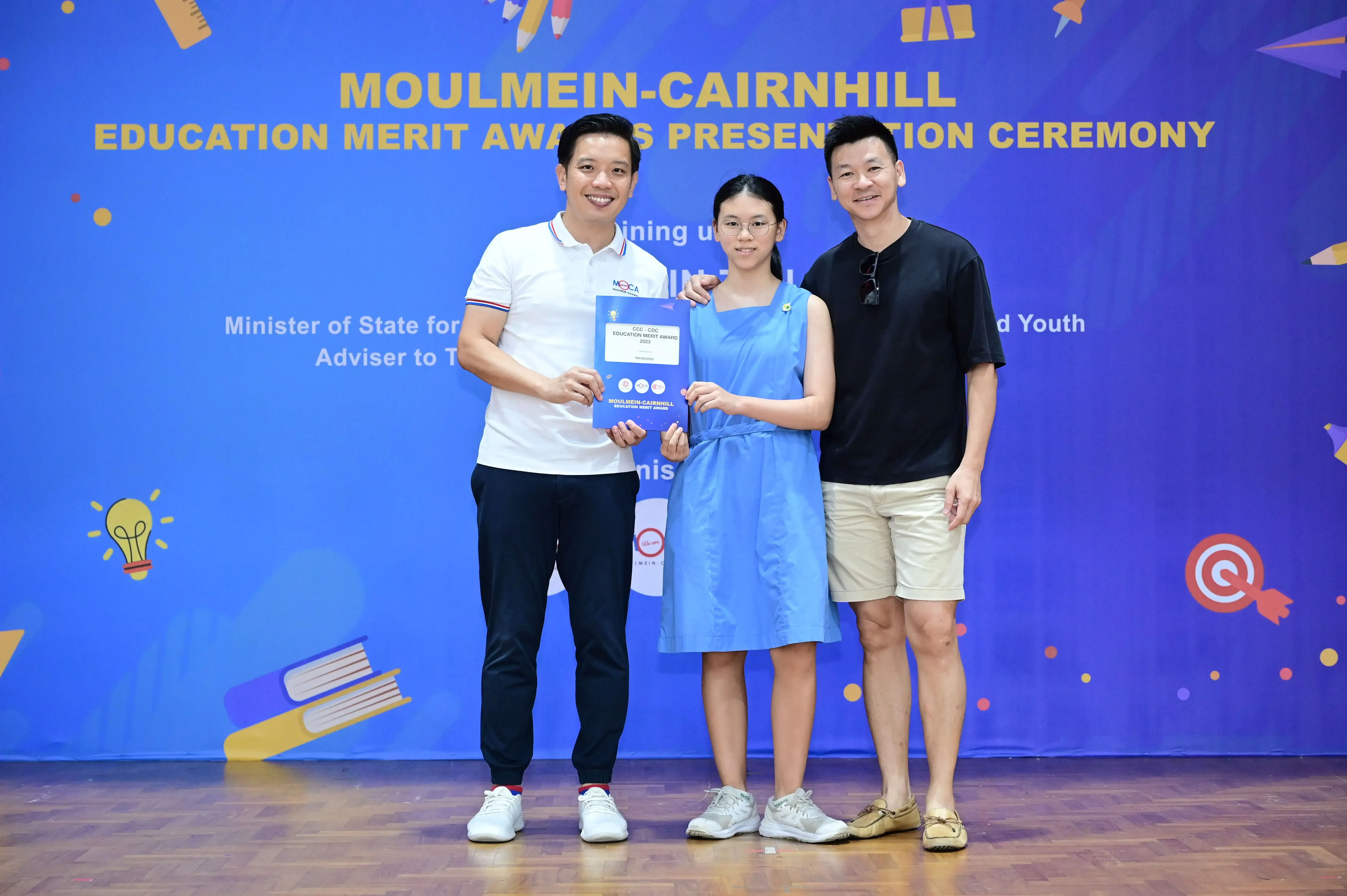 Three people pose at the Moulmein-Cairnhill Education Merit Awards ceremony, with a girl holding an award certificate.