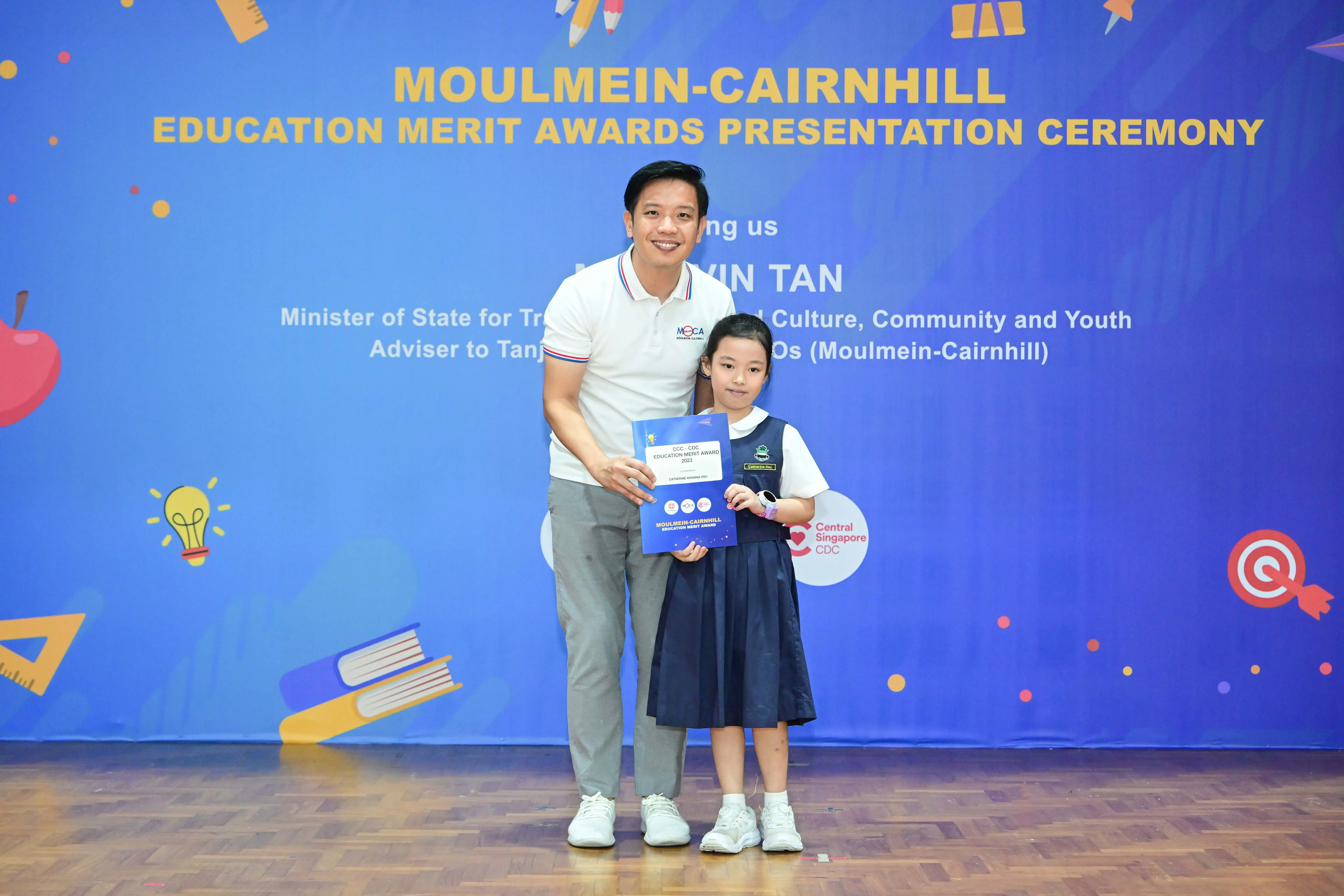 A man and a young girl in school uniform hold an award certificate at the Moulmein-Cairnhill Education Merit Awards ceremony.