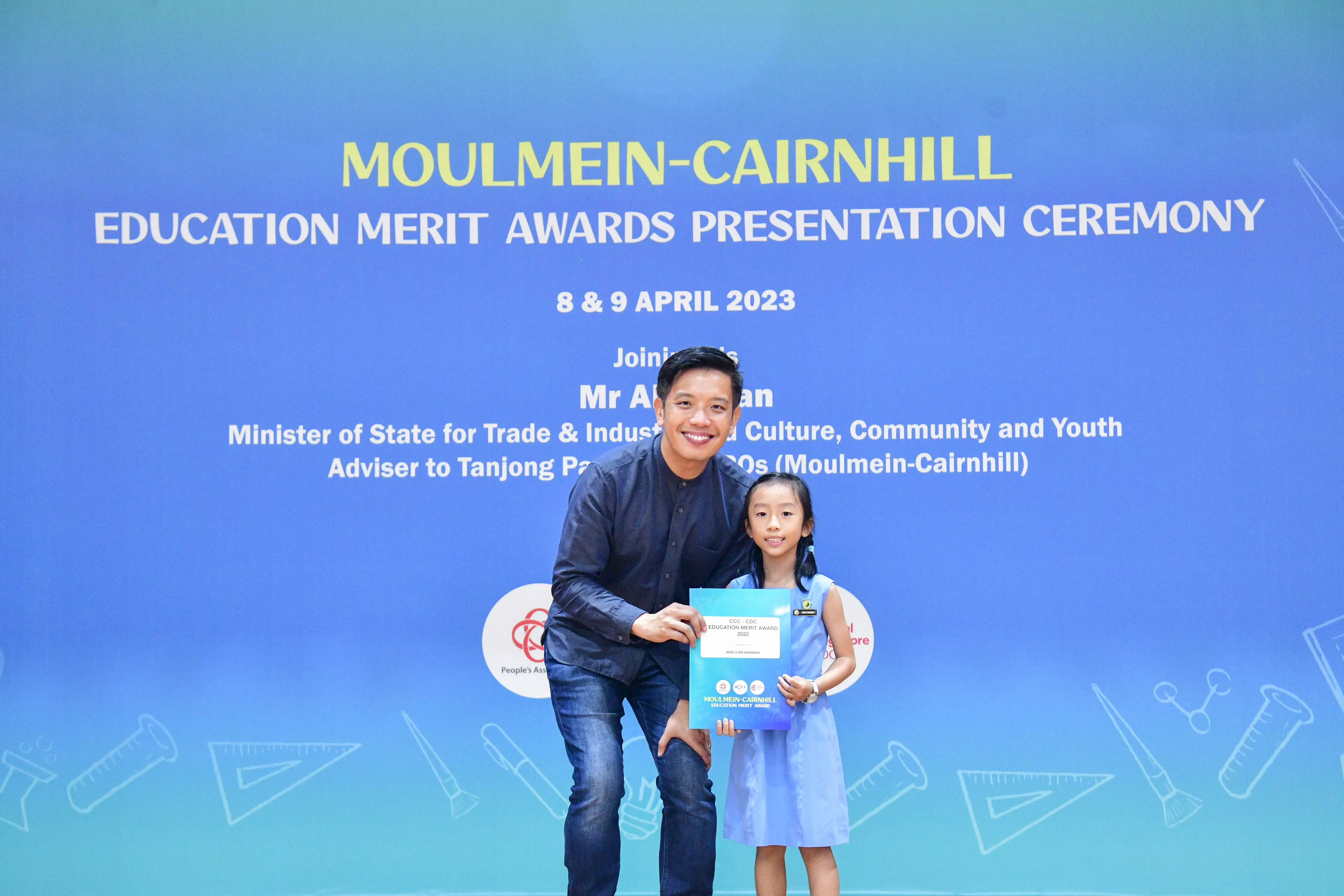 A man and a young girl pose holding an award certificate in front of a Moulmein-Cairnhill Education Merit Awards backdrop.