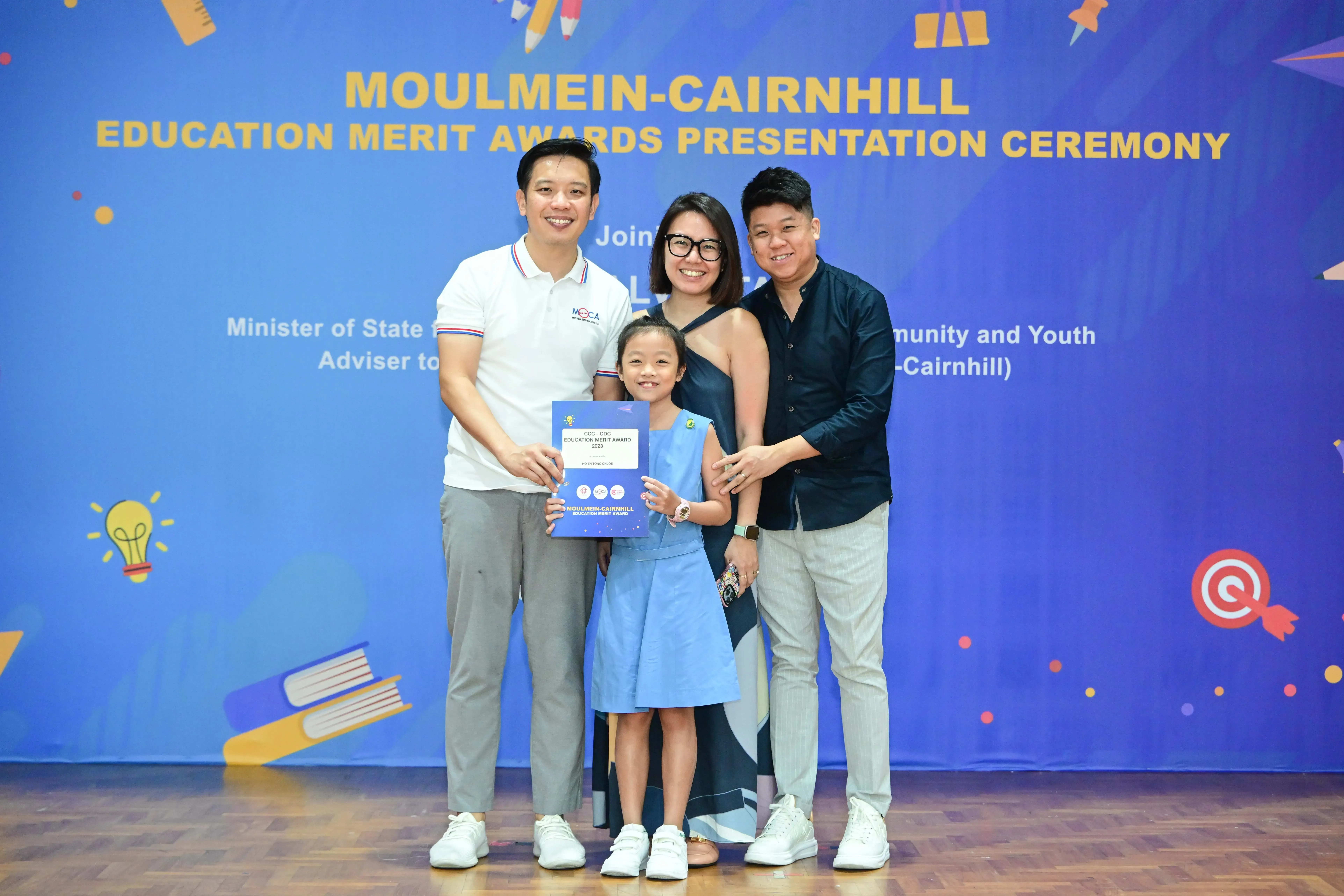 Three adults and a young girl holding an award certificate at the Moulmein-Cairnhill Education Merit Awards ceremony.