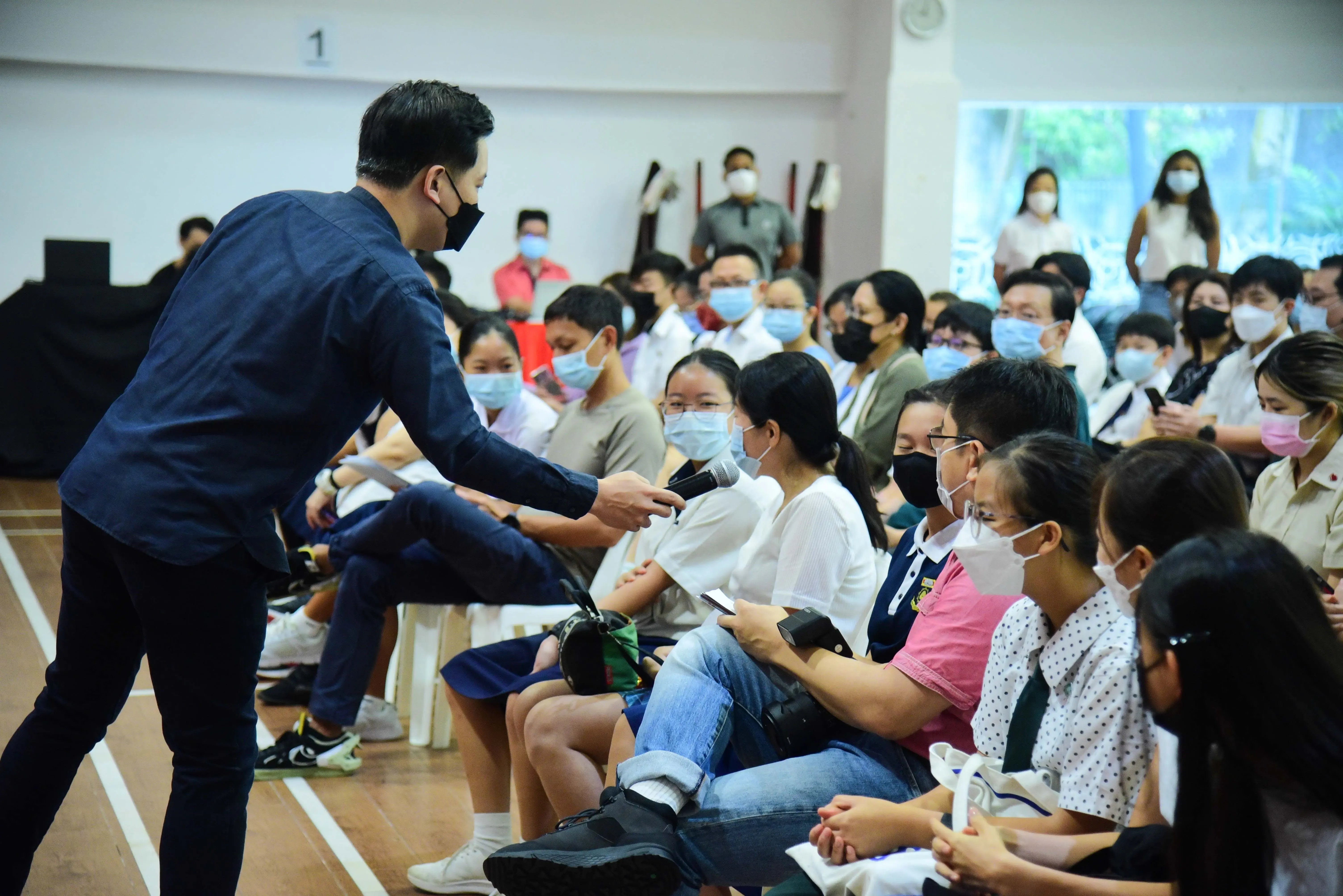 A masked man hands a microphone to a seated masked audience member in a crowded indoor hall.