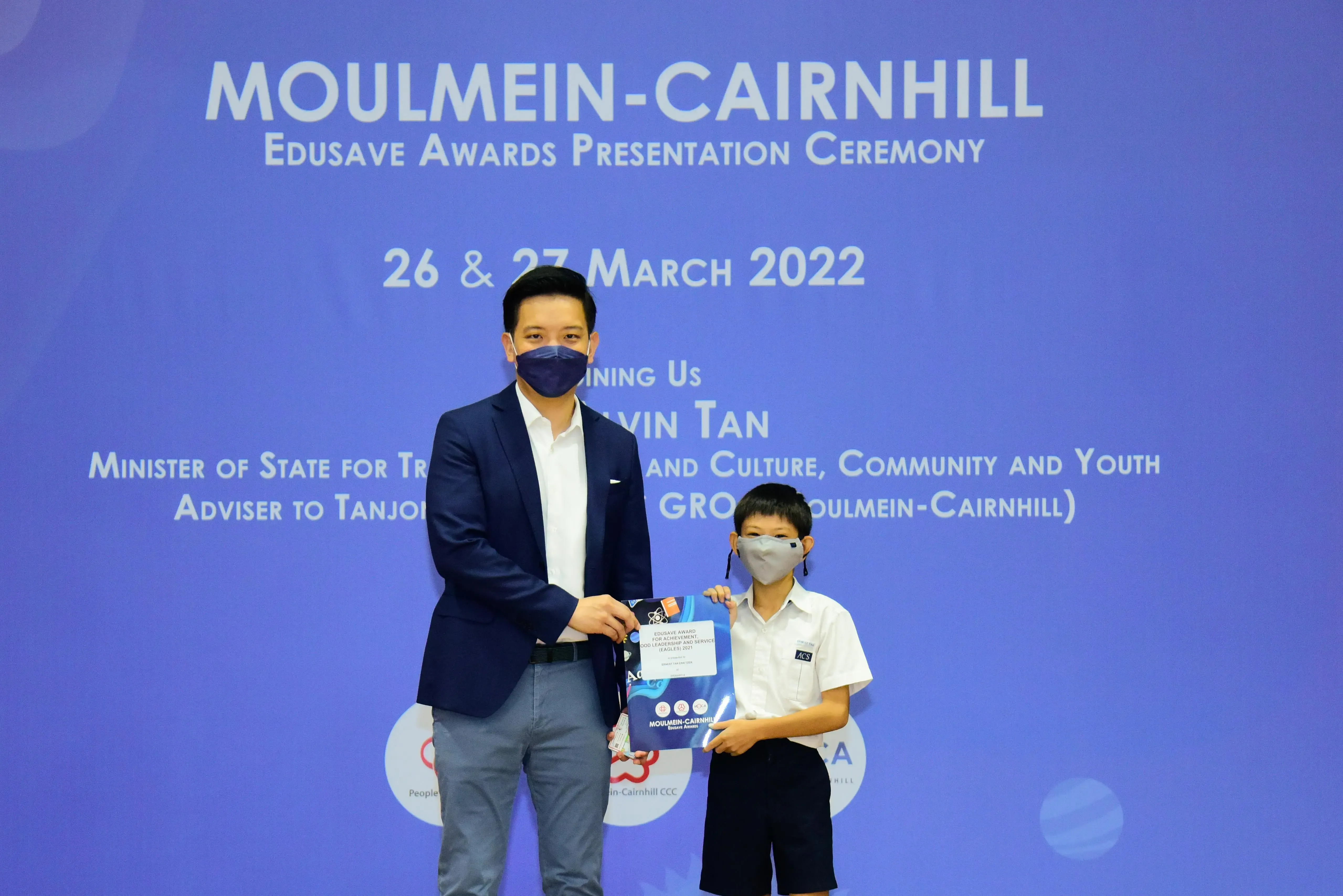 Adult in a suit and a young student in uniform hold an award at the Moulmein-Cairnhill Edusave Ceremony, March 2022.