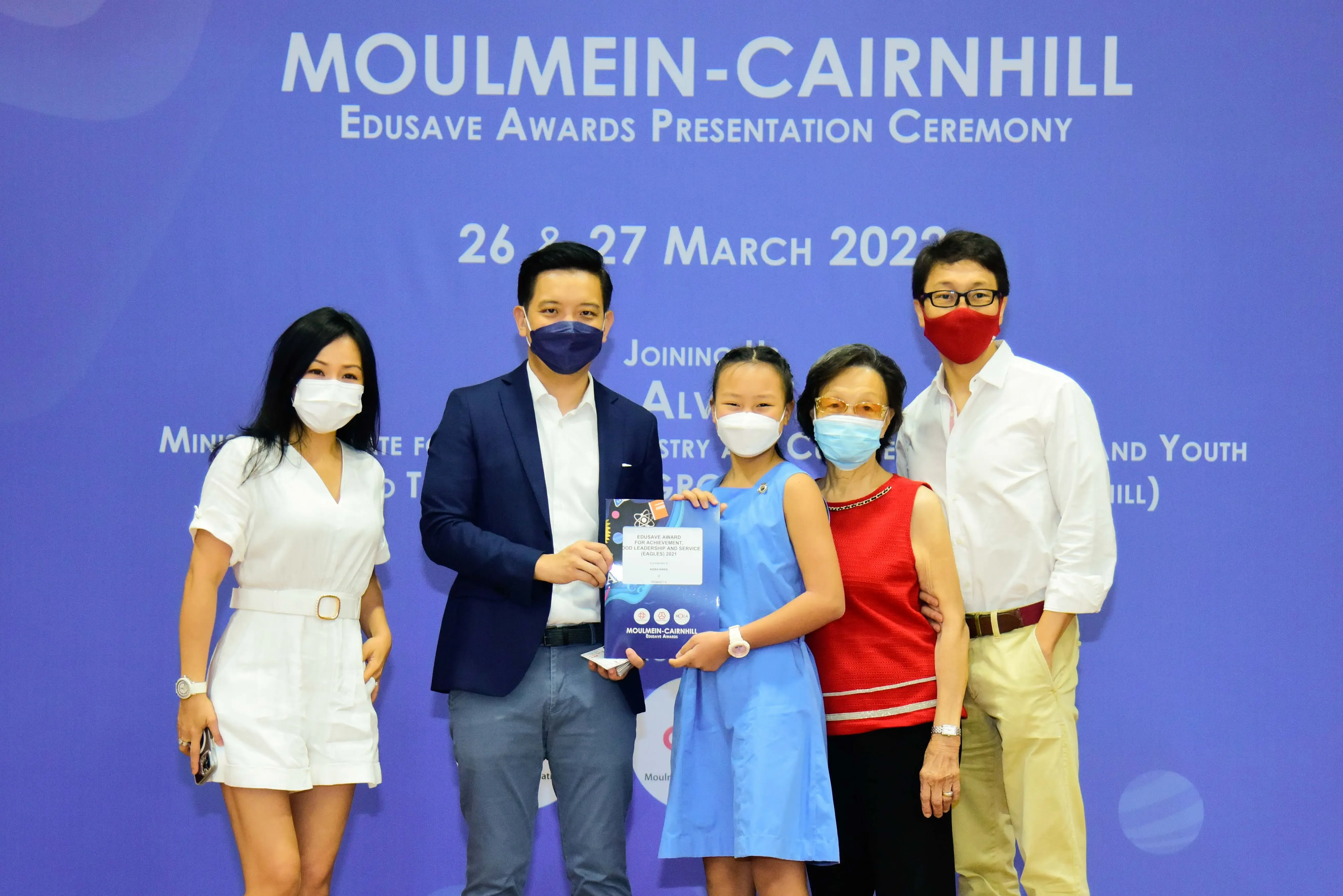Five masked people pose at the Moulmein-Cairnhill Edusave Awards Presentation Ceremony, with a child holding a certificate.