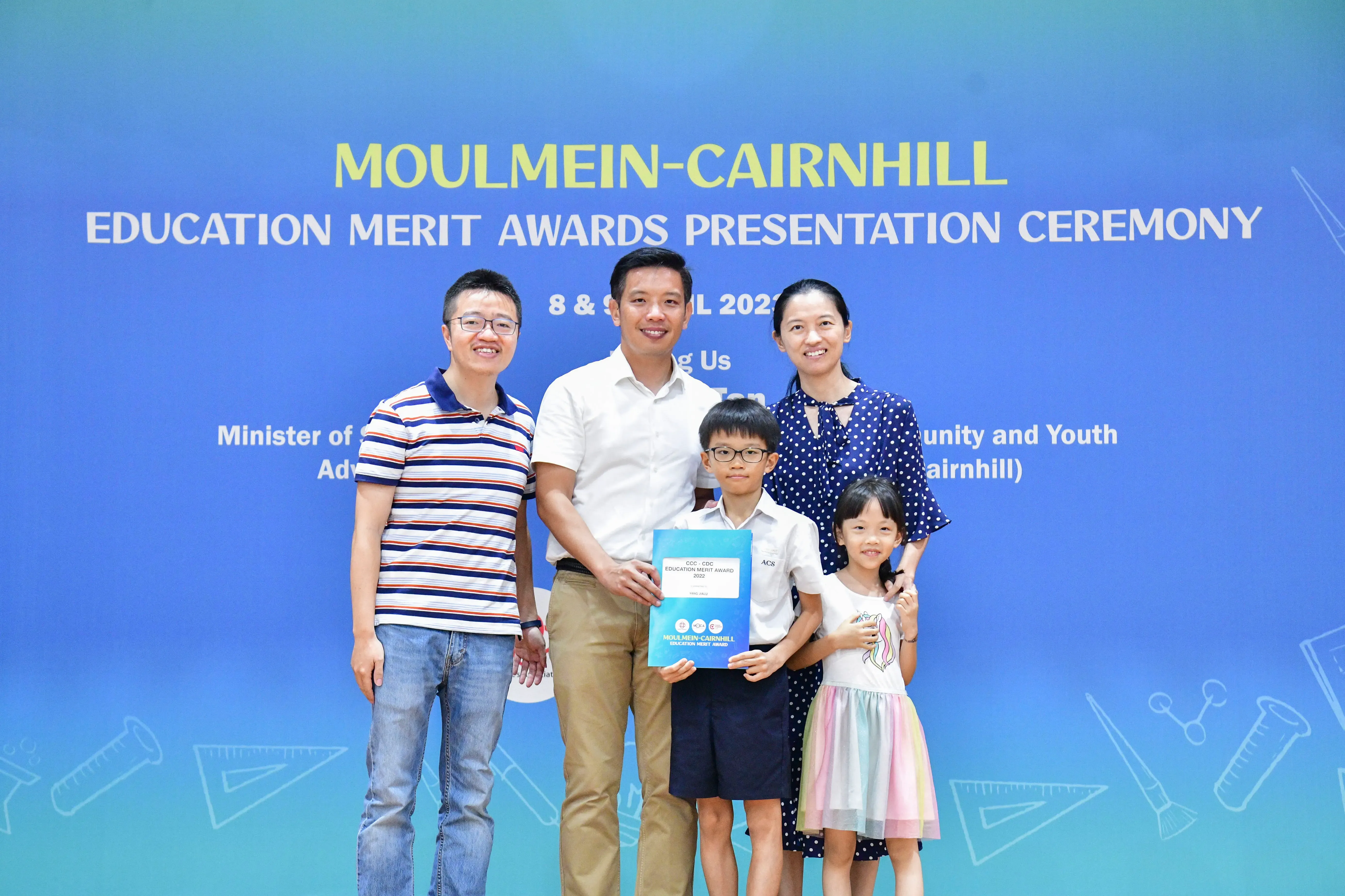 Five people pose before a Moulmein-Cairnhill Education Merit Awards banner, with a boy holding a blue award folder.