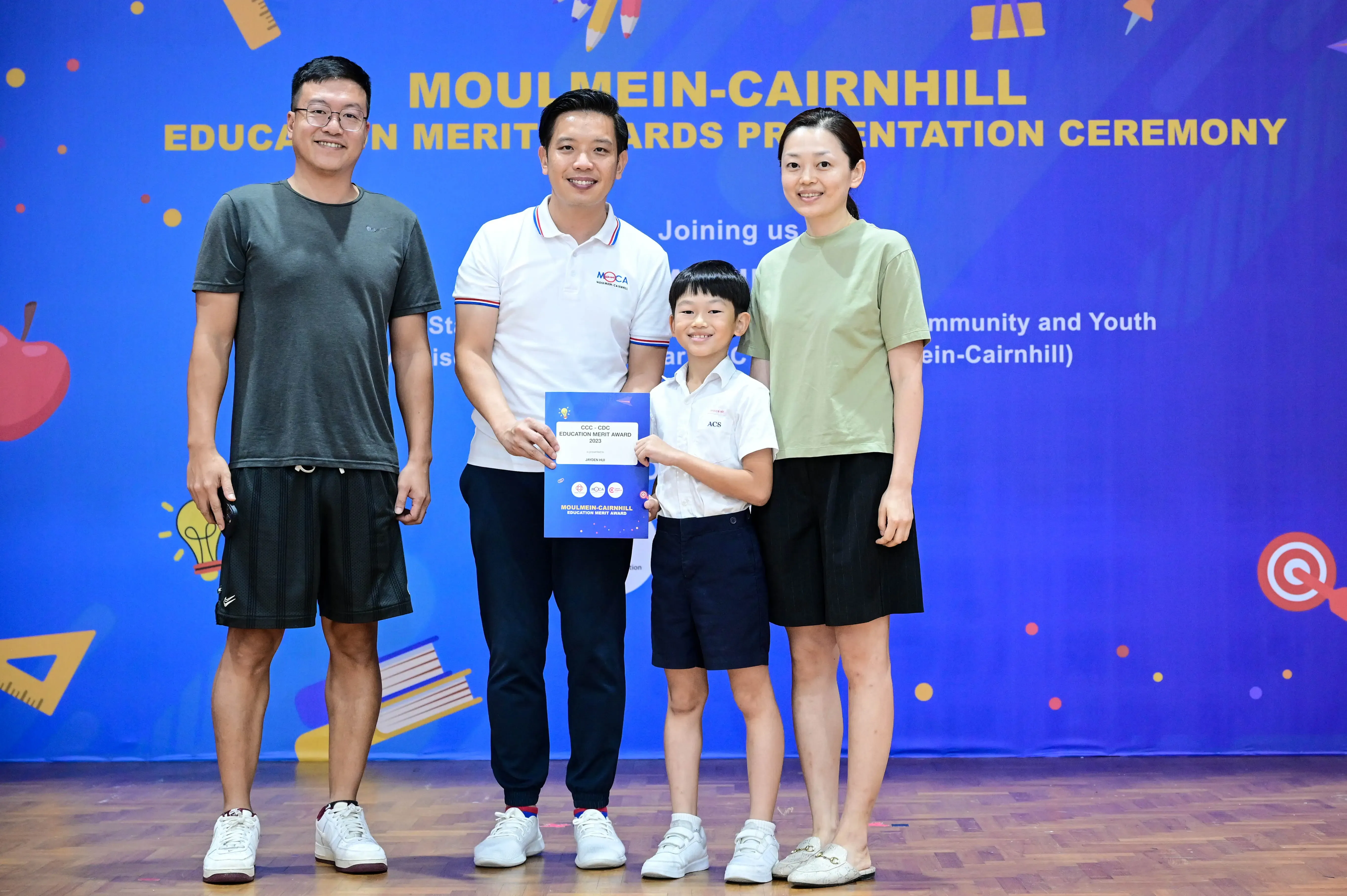 A boy holding an award certificate stands with three adults at the Moulmein-Cairnhill Education Merit Awards Ceremony.