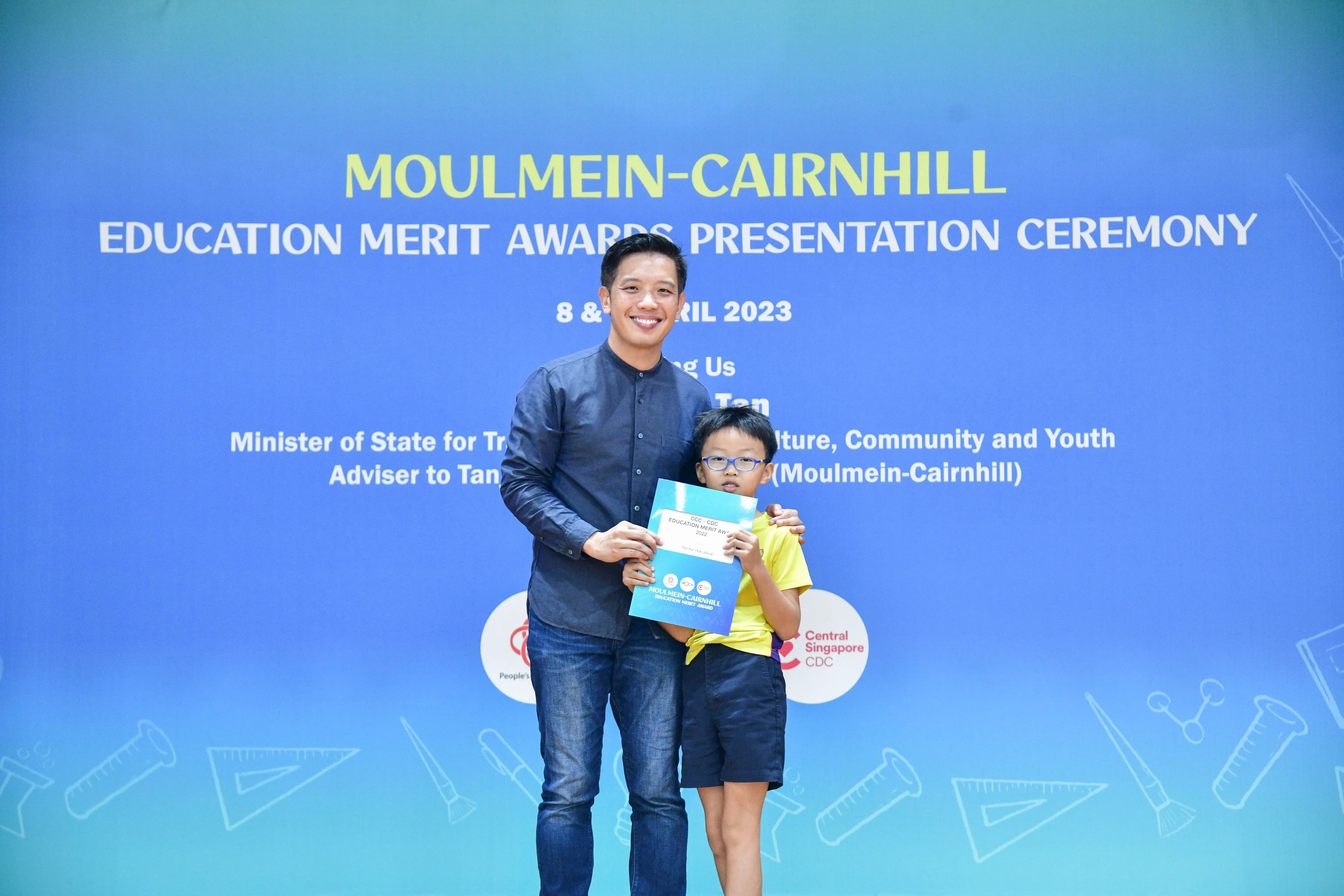 A man and a young boy holding a certificate stand before a Moulmein-Cairnhill Education Merit Awards Ceremony banner.