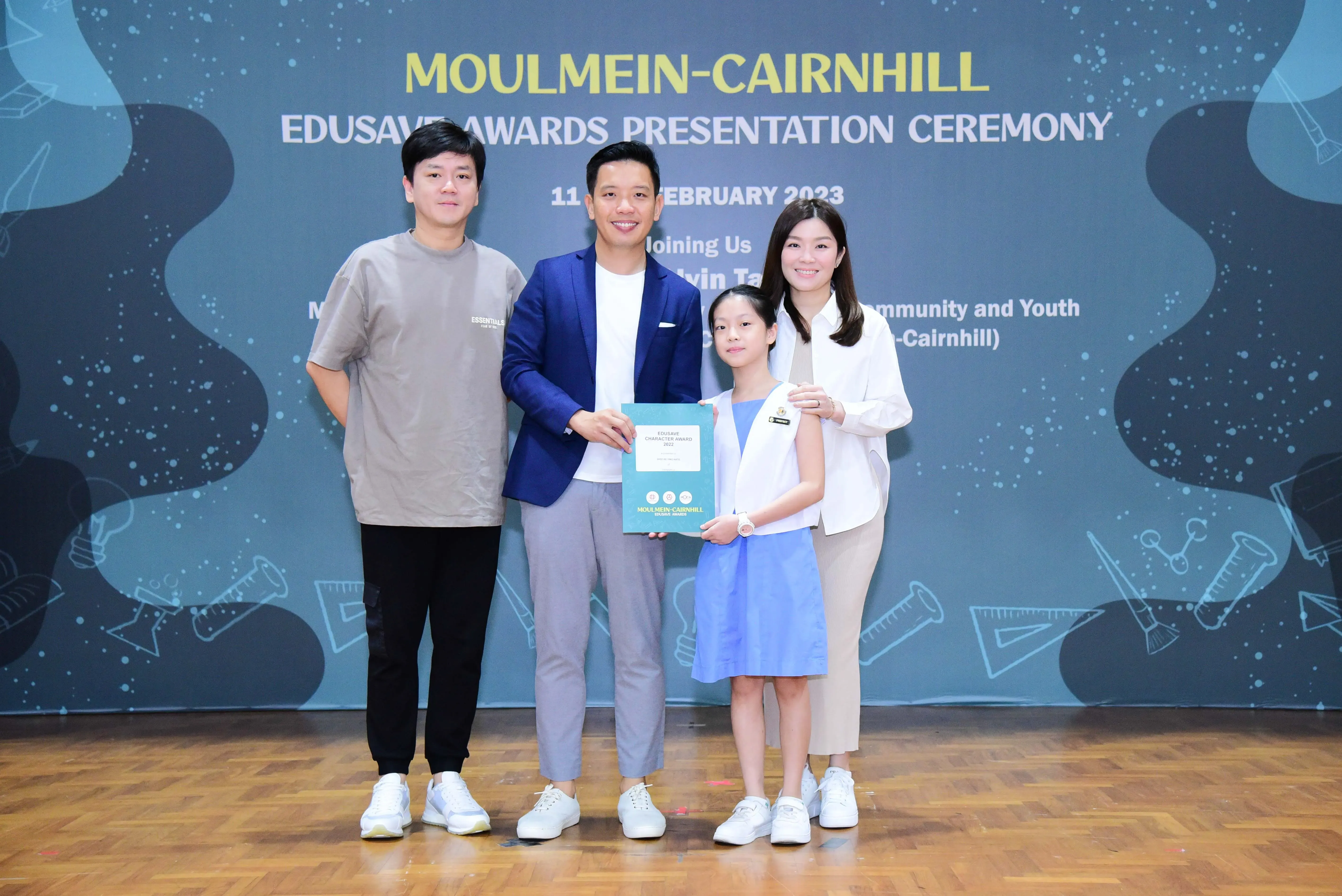 Four people pose at the Moulmein-Cairnhill Edusave Awards Presentation Ceremony, with a girl holding a certificate.