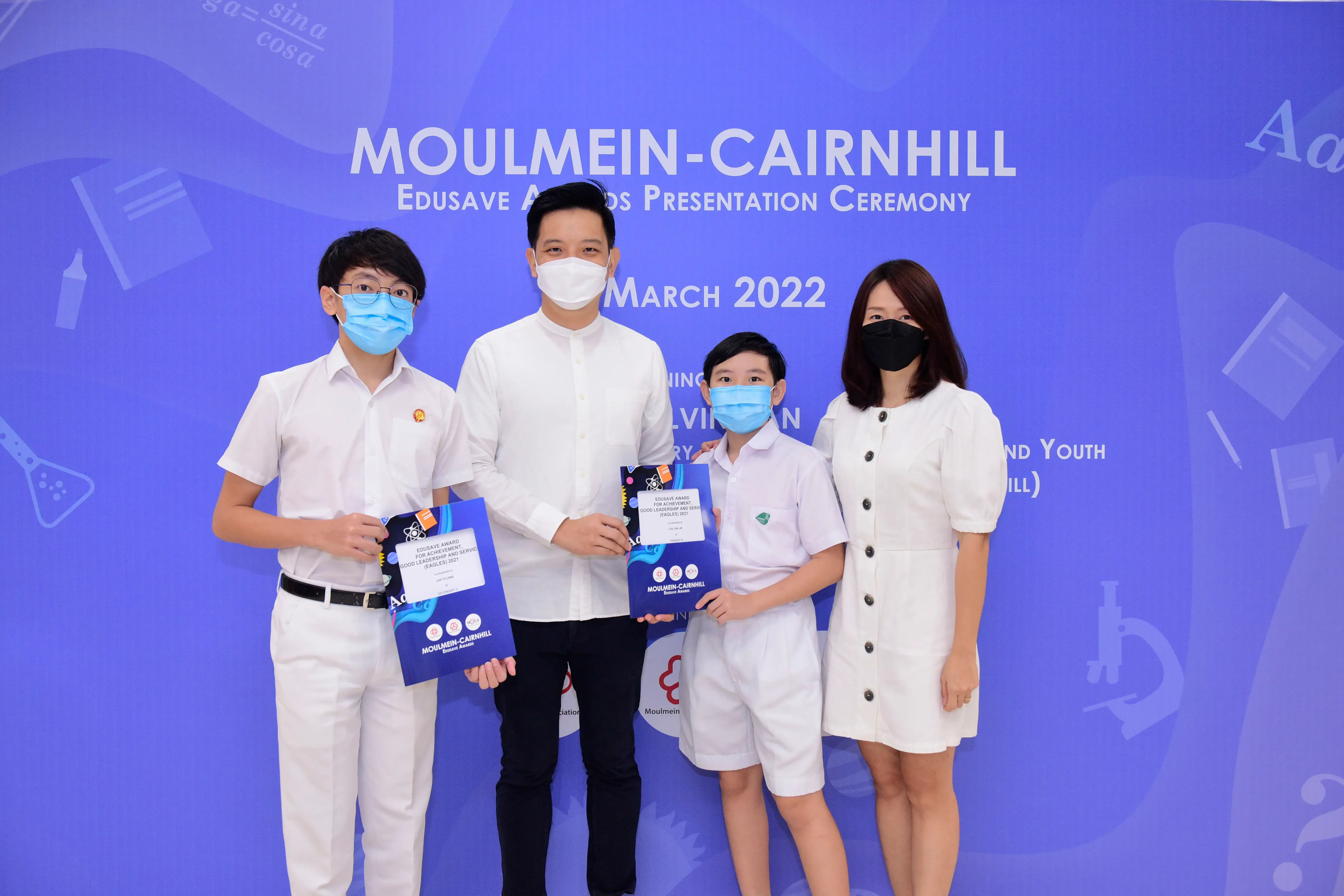 Four masked people pose at the Moulmein-Cairnhill Edusave Awards Presentation Ceremony, March 2022, two students holding certificates.