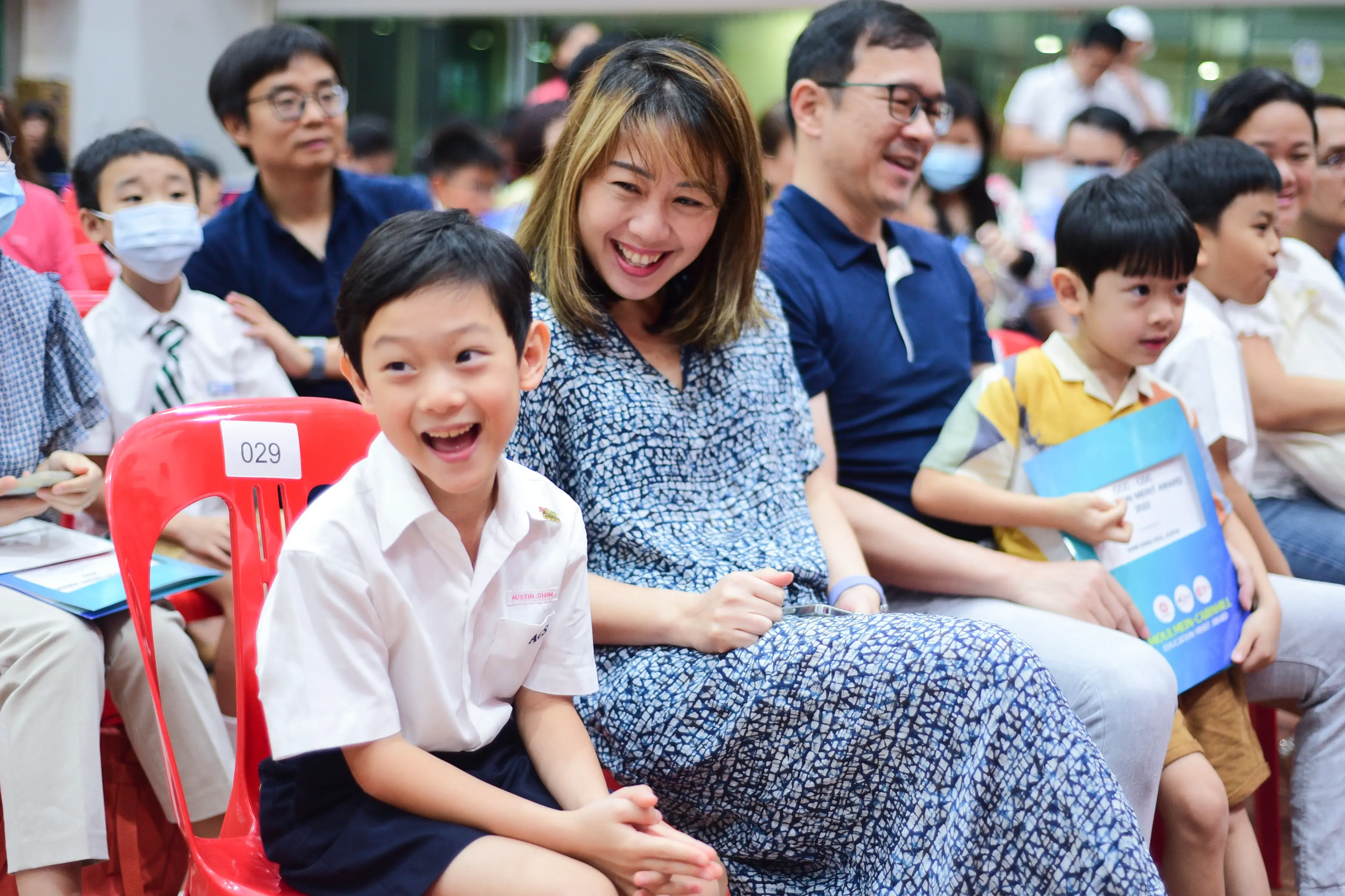 A smiling boy in school uniform sits on a red chair beside a laughing woman at a crowded school event.