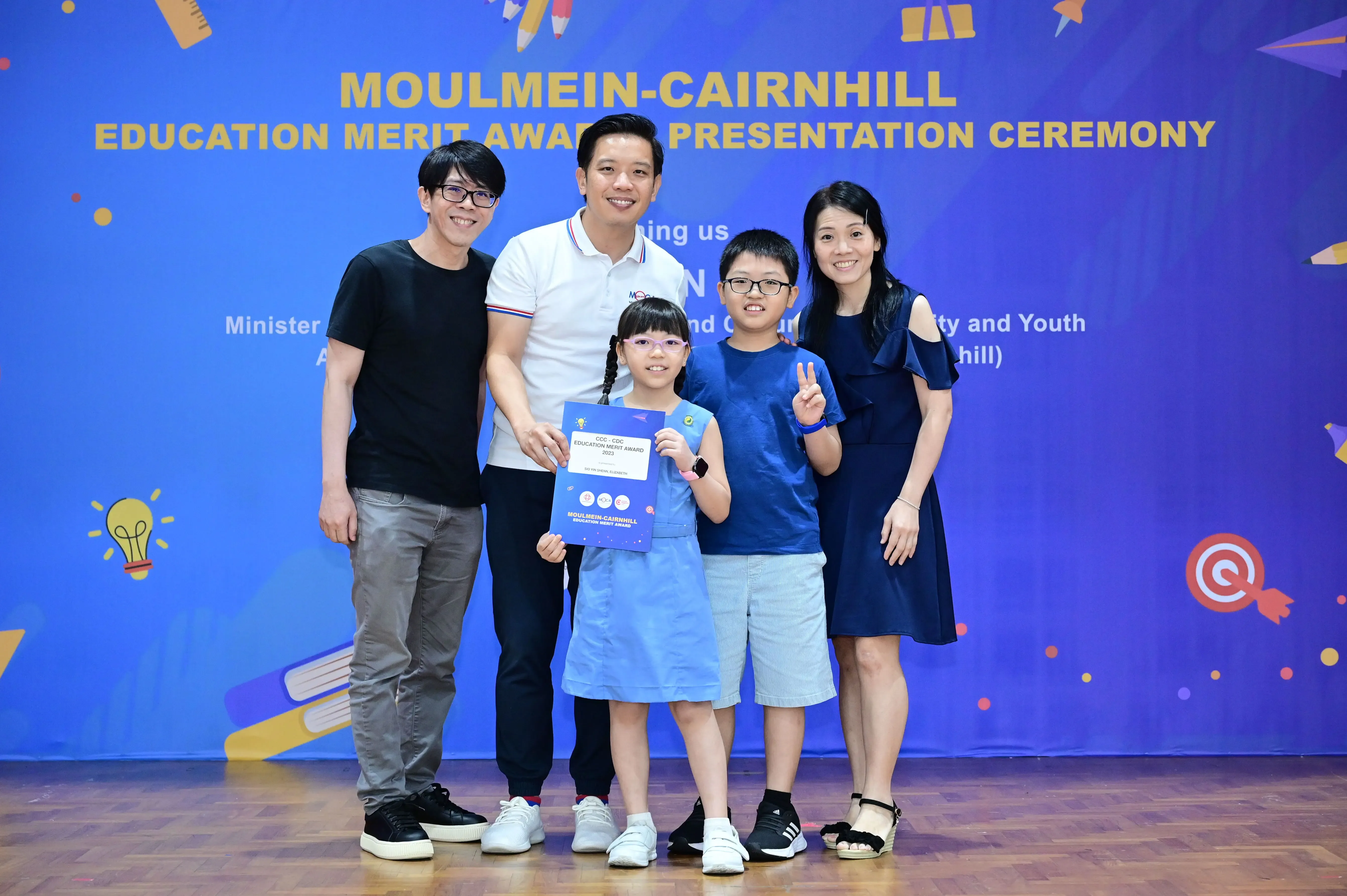 Five people pose smiling before a Moulmein-Cairnhill Education Merit Award Presentation Ceremony banner.