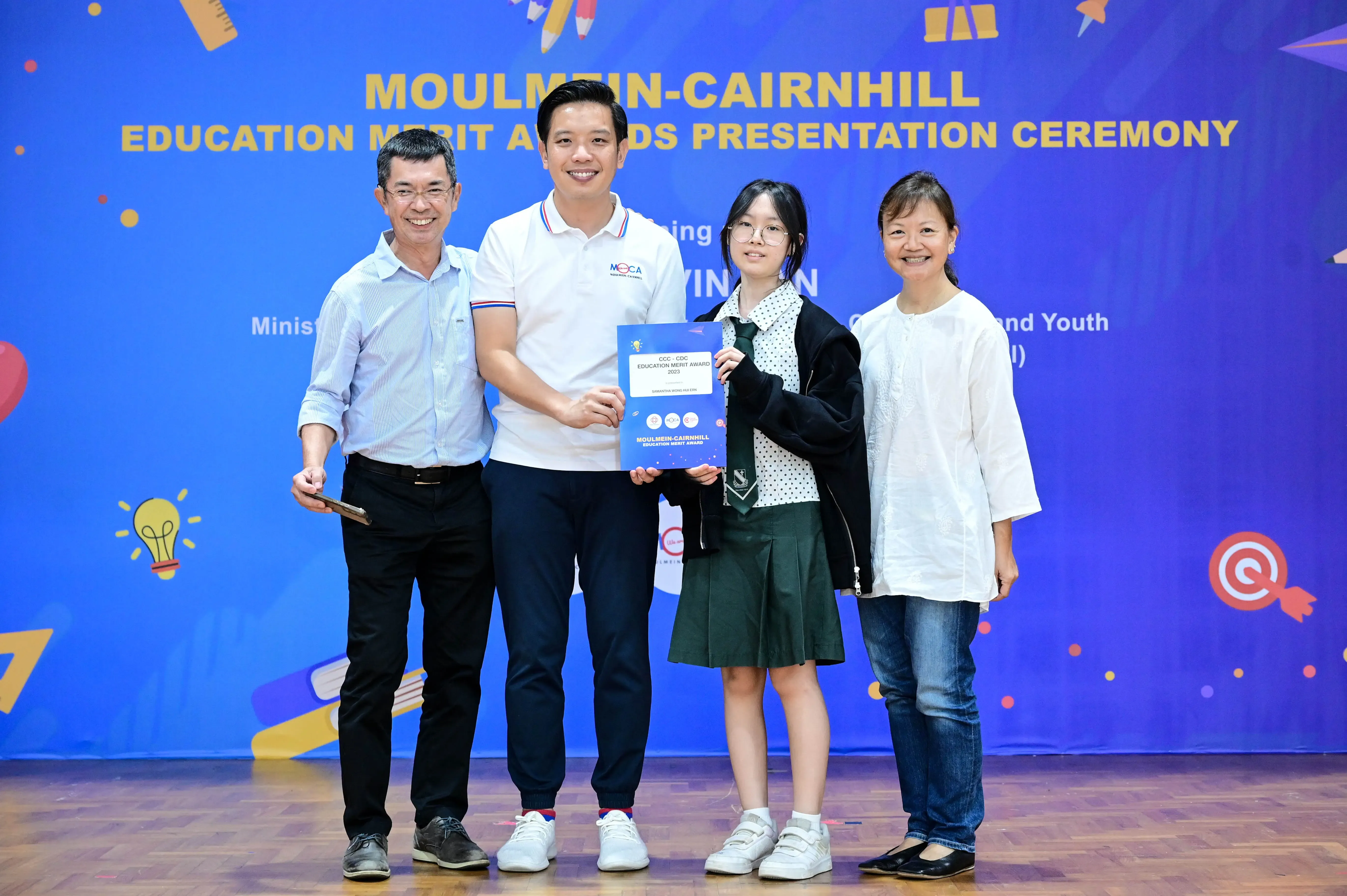 Four people pose on stage at the Moulmein-Cairnhill Education Merit Awards Presentation Ceremony, holding a certificate.