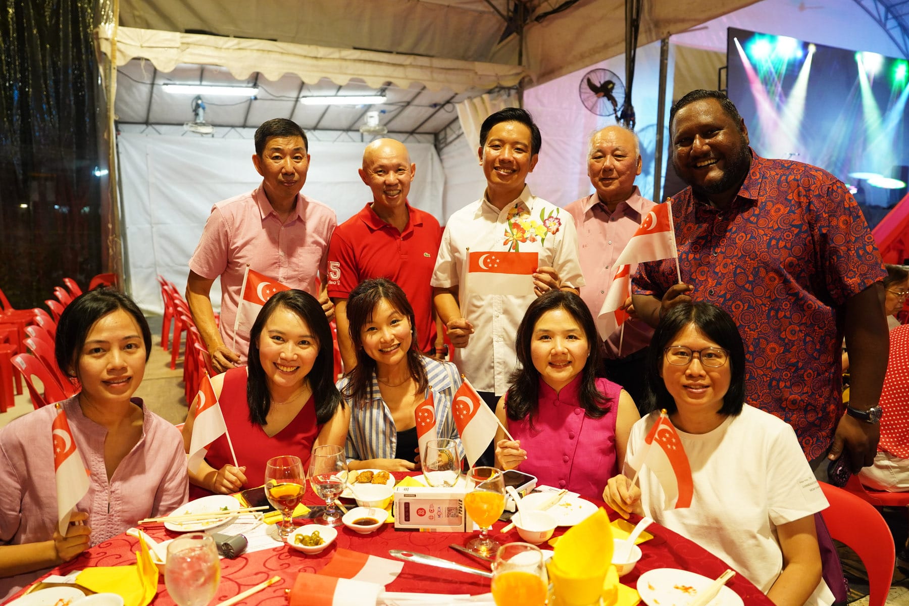 A group of nine smiling people holding Singapore flags at an outdoor dinner event with food and drinks on a red-clothed table.